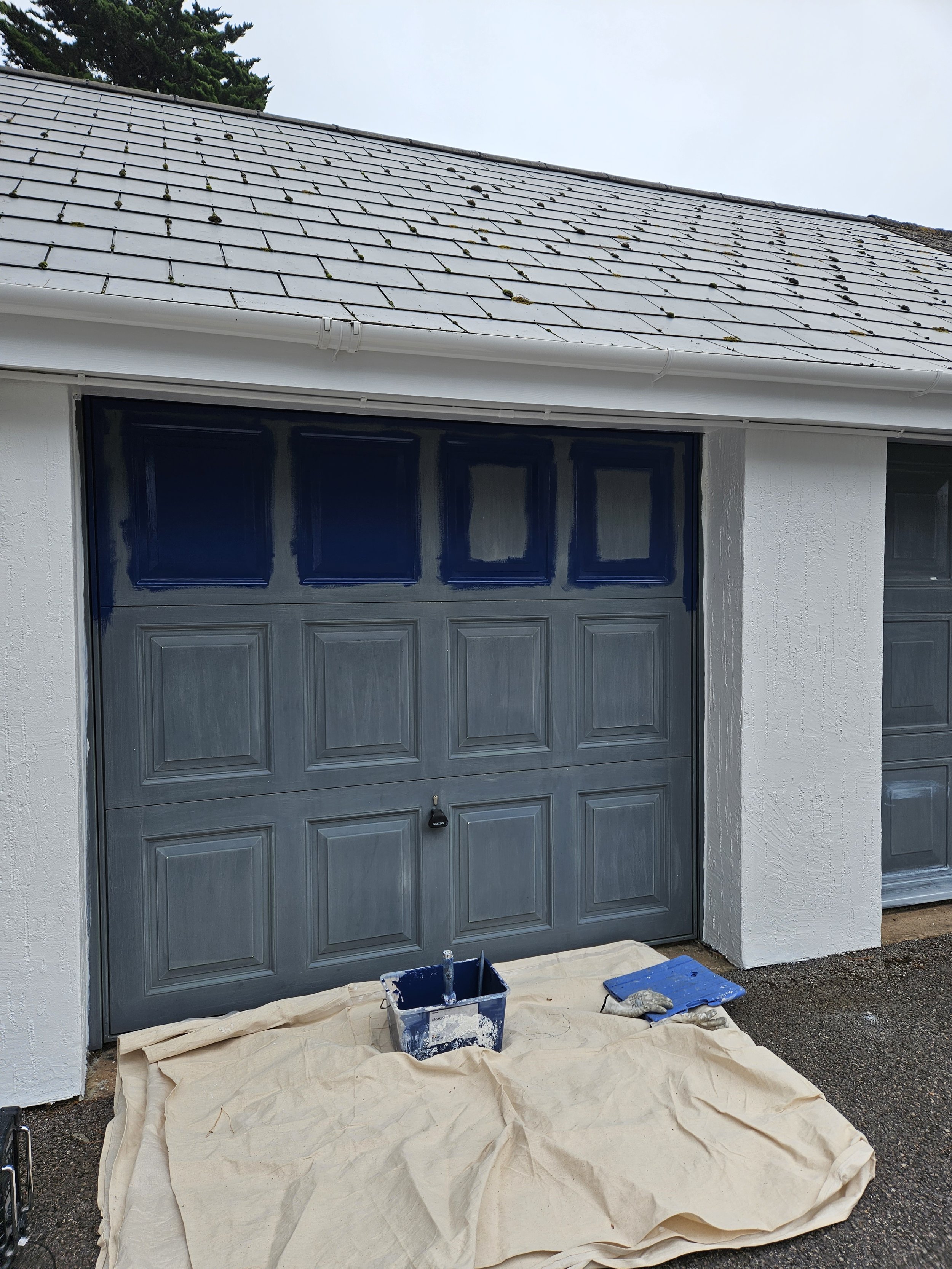 Garage door painting in progress with paint bucket, roller, and tarp on the ground, in front of a white textured wall and house roof.