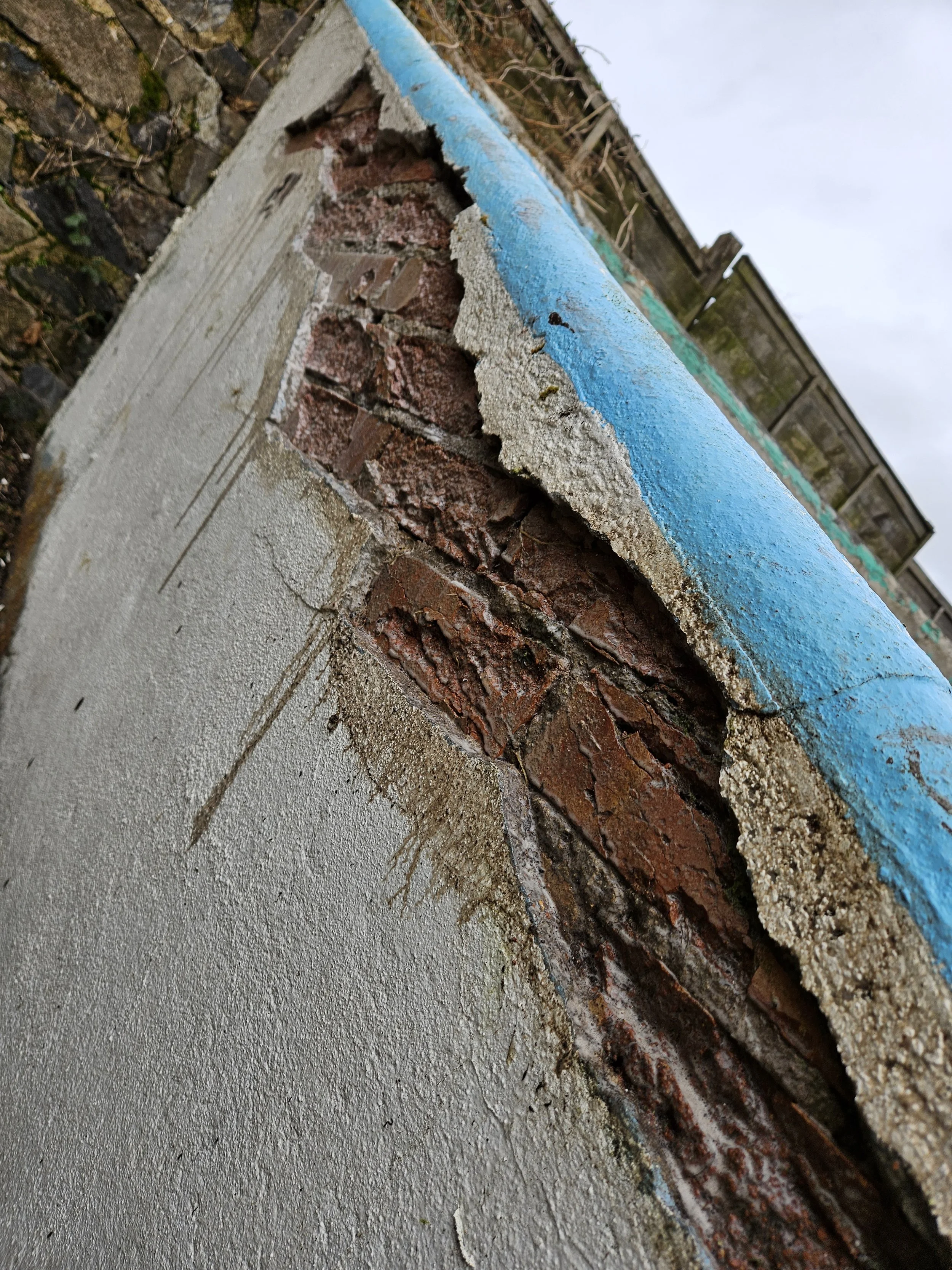 Close-up of a wall with peeling blue paint, exposing red bricks and gray concrete underneath, outdoors.