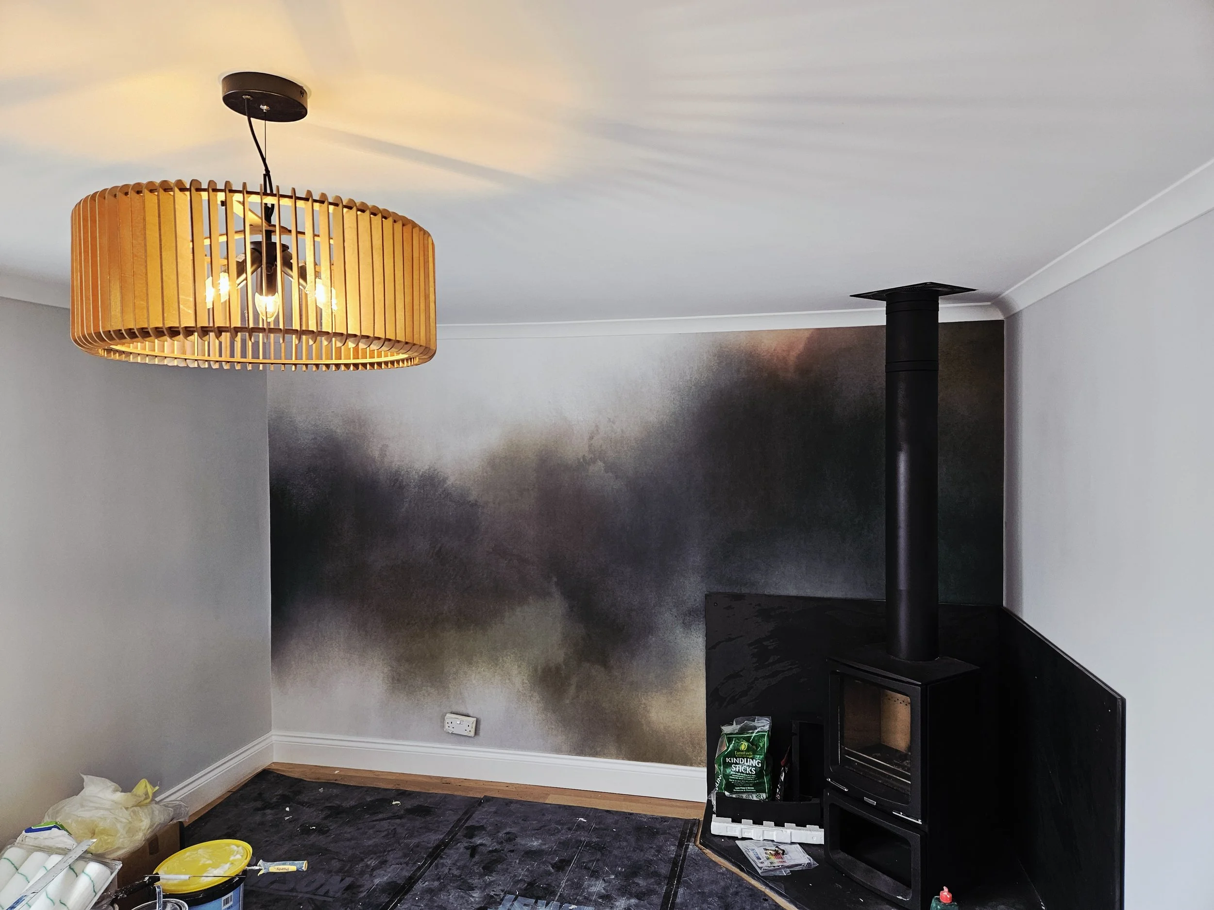 A living room with a black stove, a large black pipe, a decorative wall, and a modern light fixture with a wooden frame hanging from the ceiling.