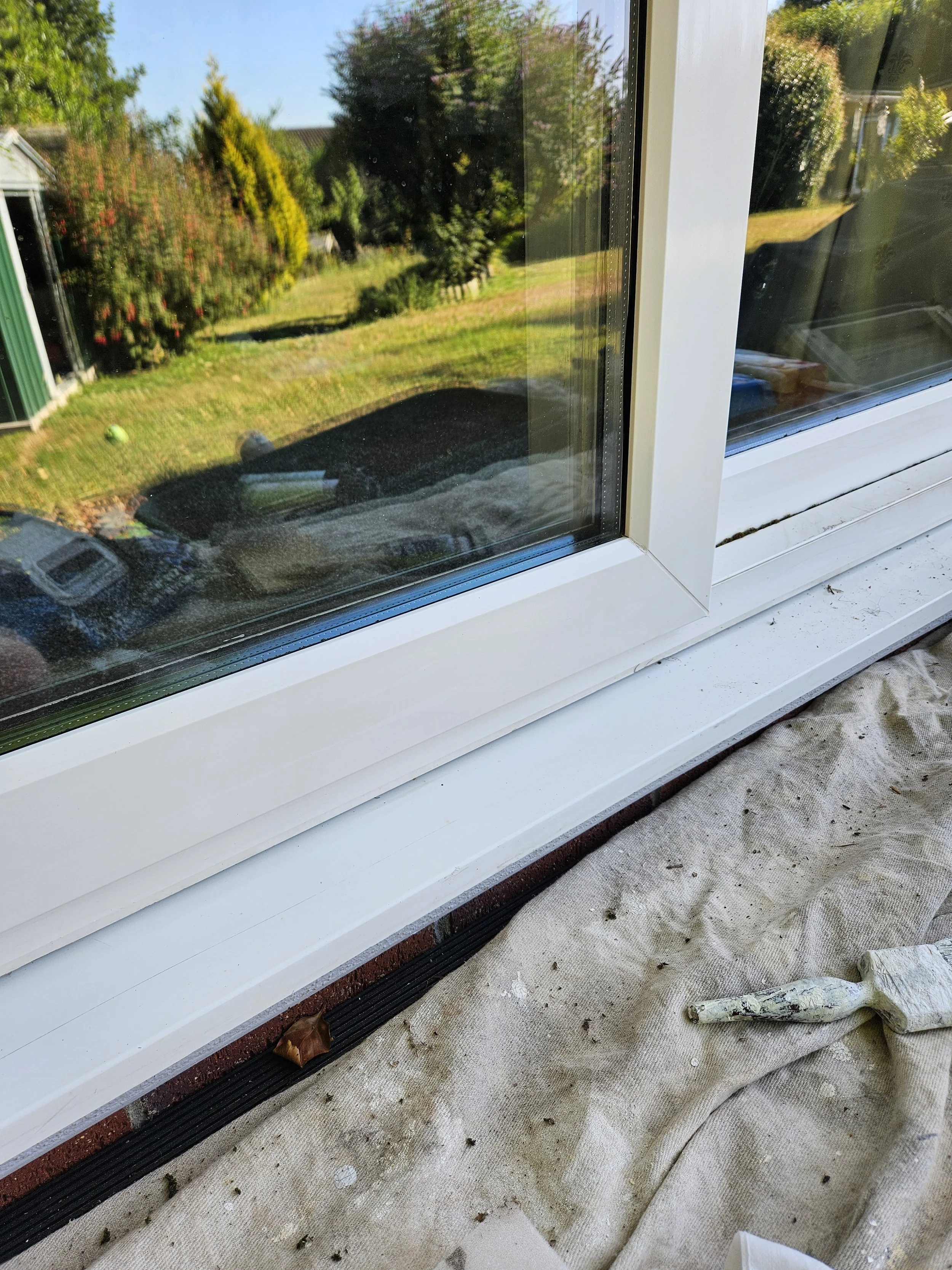 Close-up of a white window frame with a garden view outside, showing trees and a grassy yard. The window is part of a construction or renovation project, with a cloth covering the windowsill and a construction tool or tube lying on the cloth.