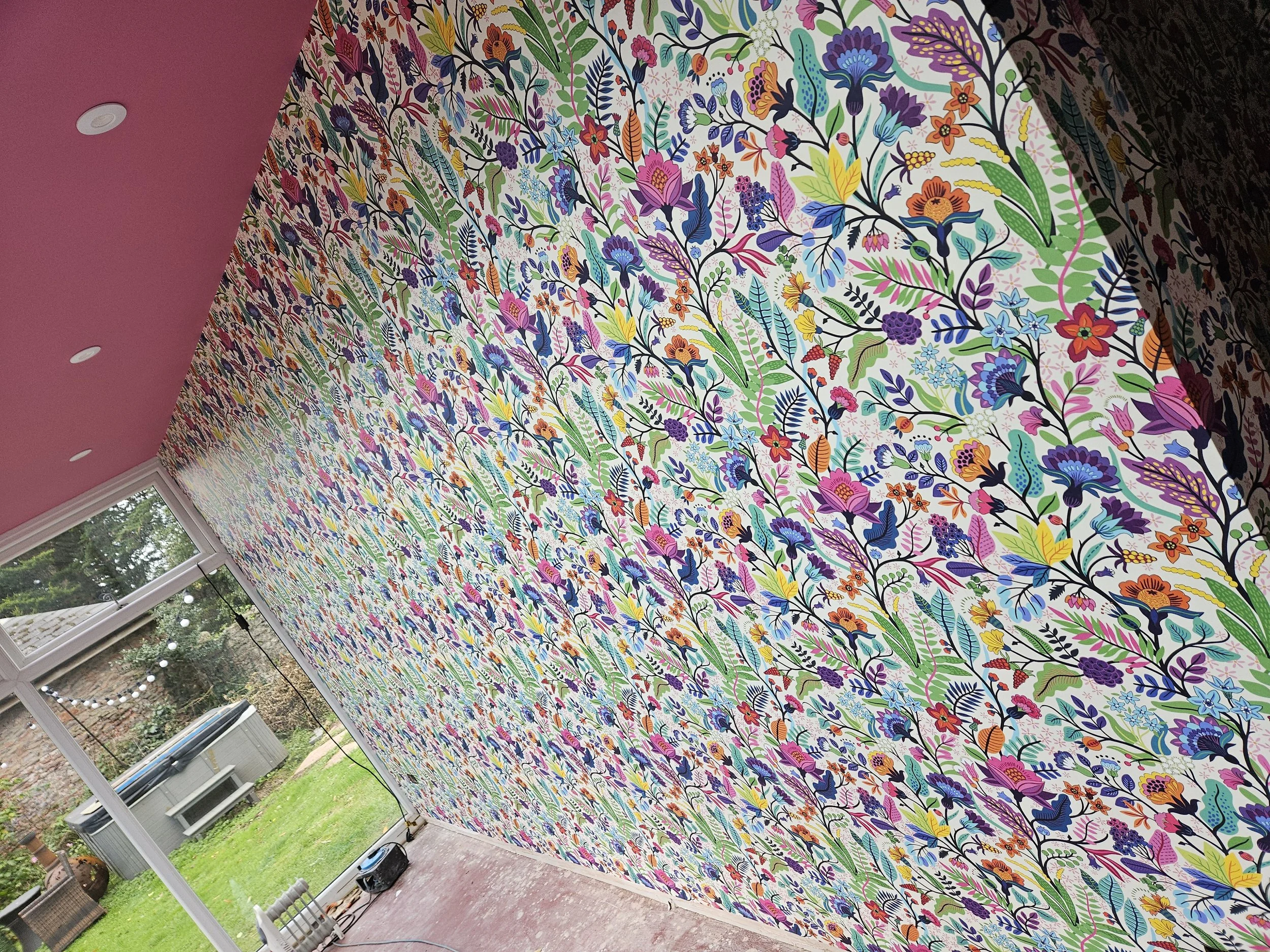 A room with a wall covered in colorful floral wallpaper. The room has a pink ceiling with recessed lighting. There is a glass door leading to an outdoor area with grass and trees, visible through the door. An air conditioning unit and some outdoor fu