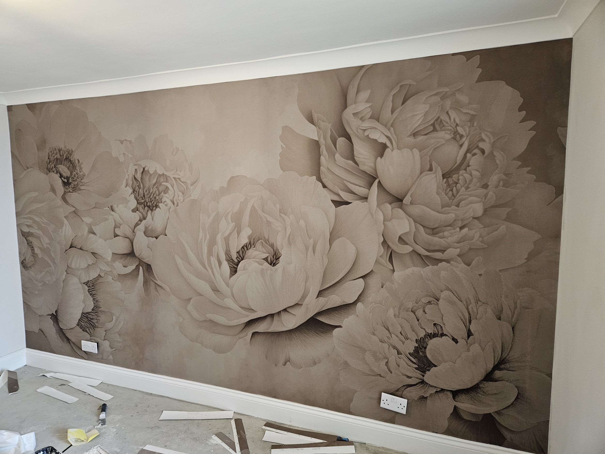 A large floral mural with detailed peony flowers in soft, neutral shades covers the wall of a room under construction, with electrical outlets visible near the floor.