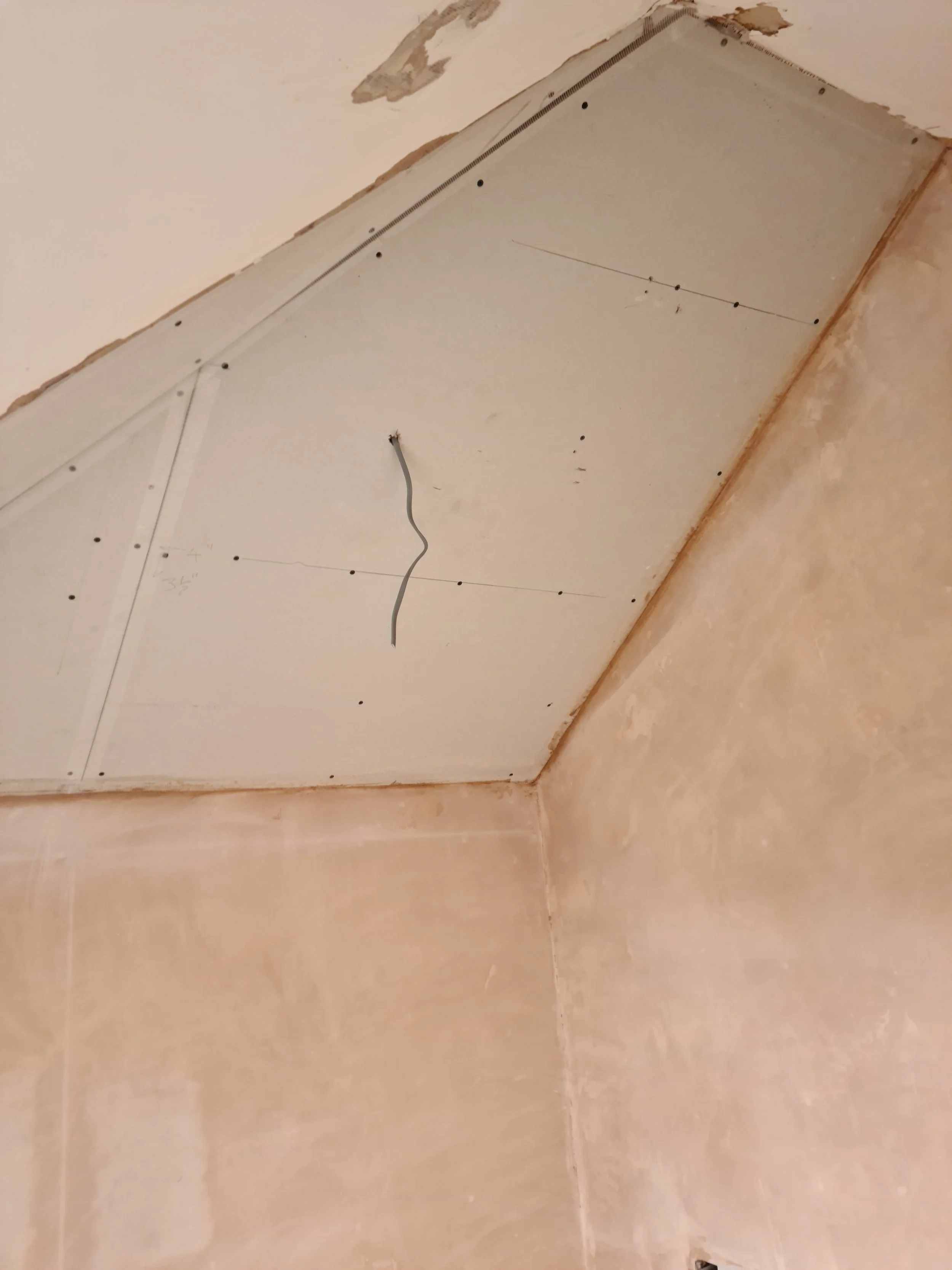 Ceiling with drywall panels partially installed, with a hole and a wire hanging down, surrounded by unpainted wall surfaces.