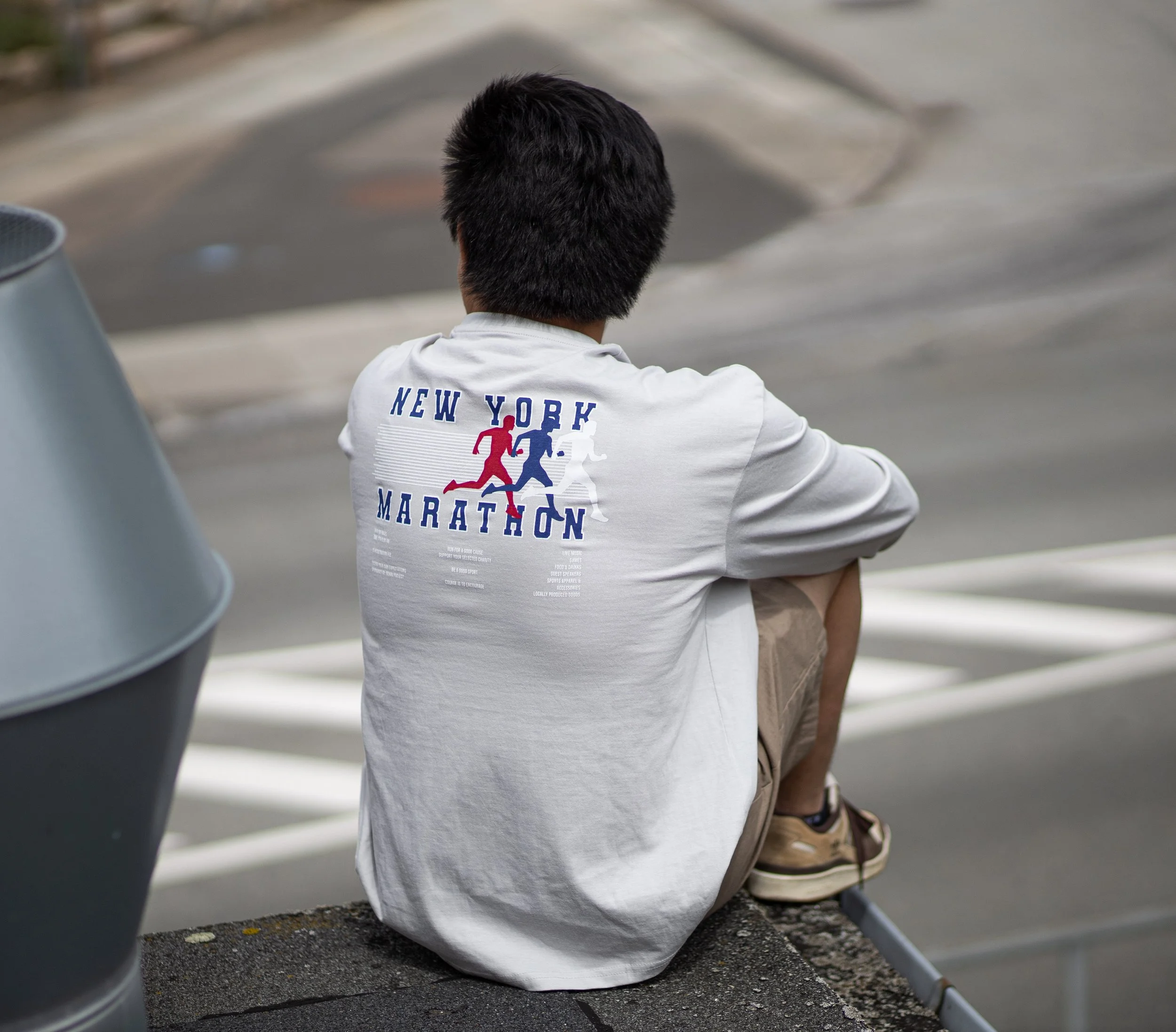 En person sidder på en kantsten med ryggen til, iført en hvid T-shirt med teksten 'New York Marathon' og en grafik af to løbere på. Personen har mørkt, kort hår og bærer sandaler og shorts.