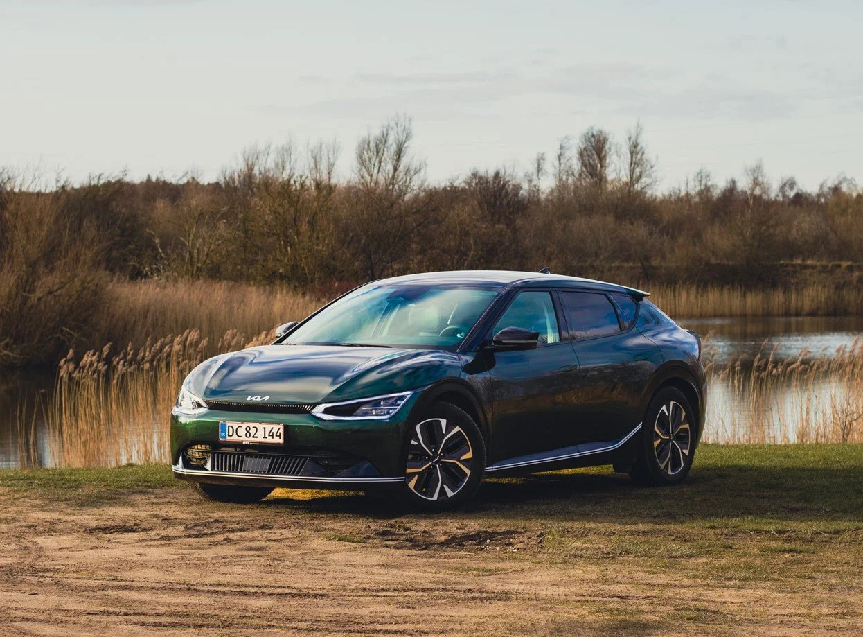 En sort elektrisk Kia bil parkeret på en græsmark ved en sø med siv langs bredden, under en overskyet himmel.