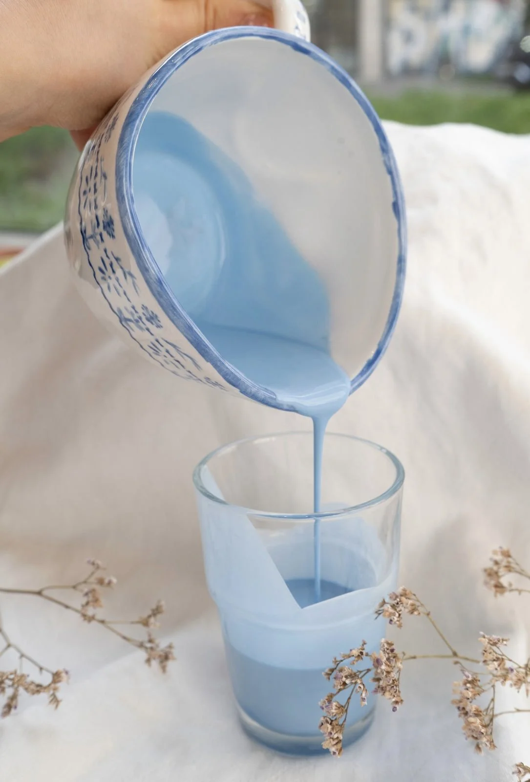 Blaue Farbe wird aus einer Pappschale in ein Glas gegossen, auf einem hellen Tuch mit kleinen Trockenblumen, im Freien.