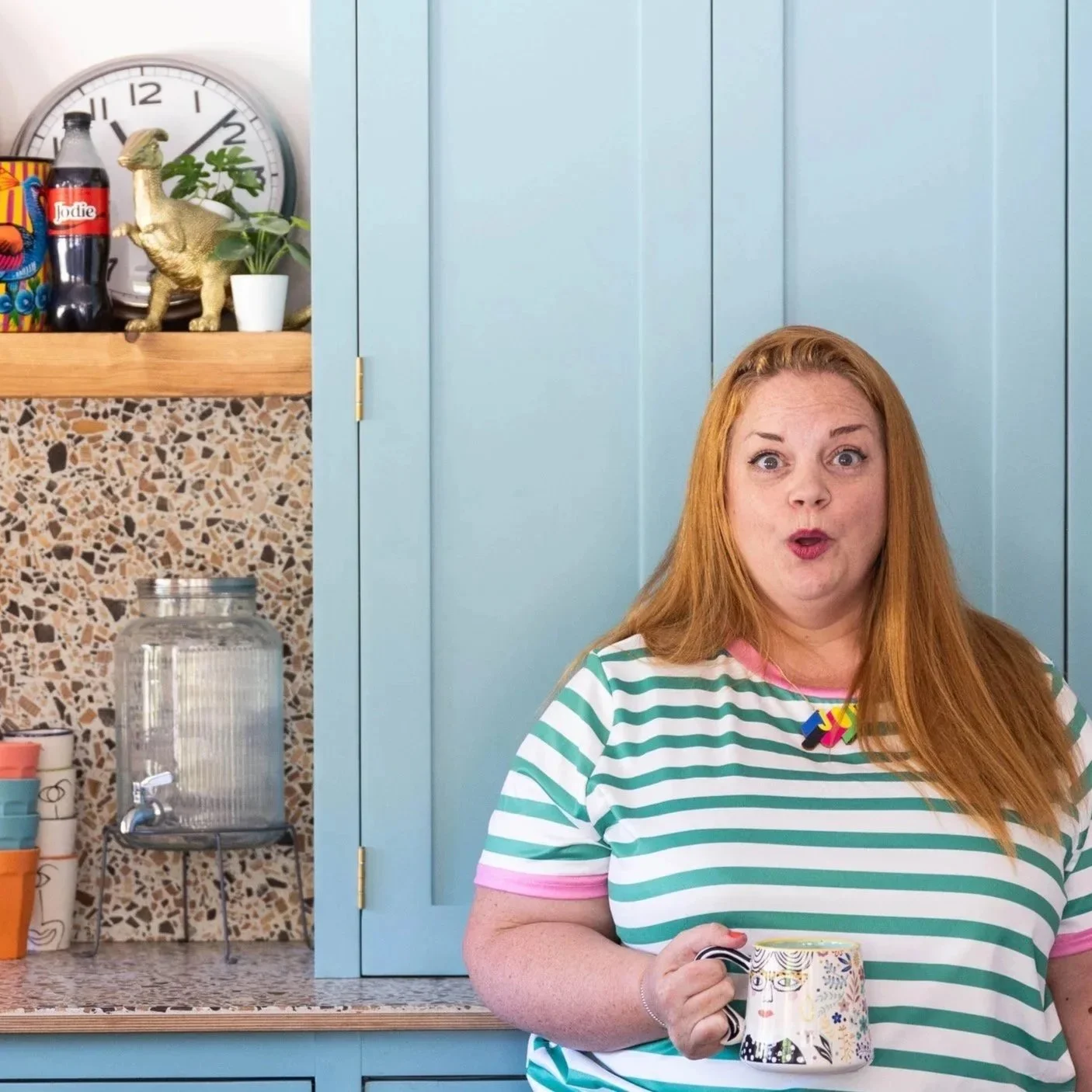 A woman with red hair and a striped shirt holding a mug in a kitchen with light blue cabinets, a water dispenser with a tap, and tiled backsplash. There are decorative items on the shelves, including a dinosaur figure, a clock, plants, and colorful containers.