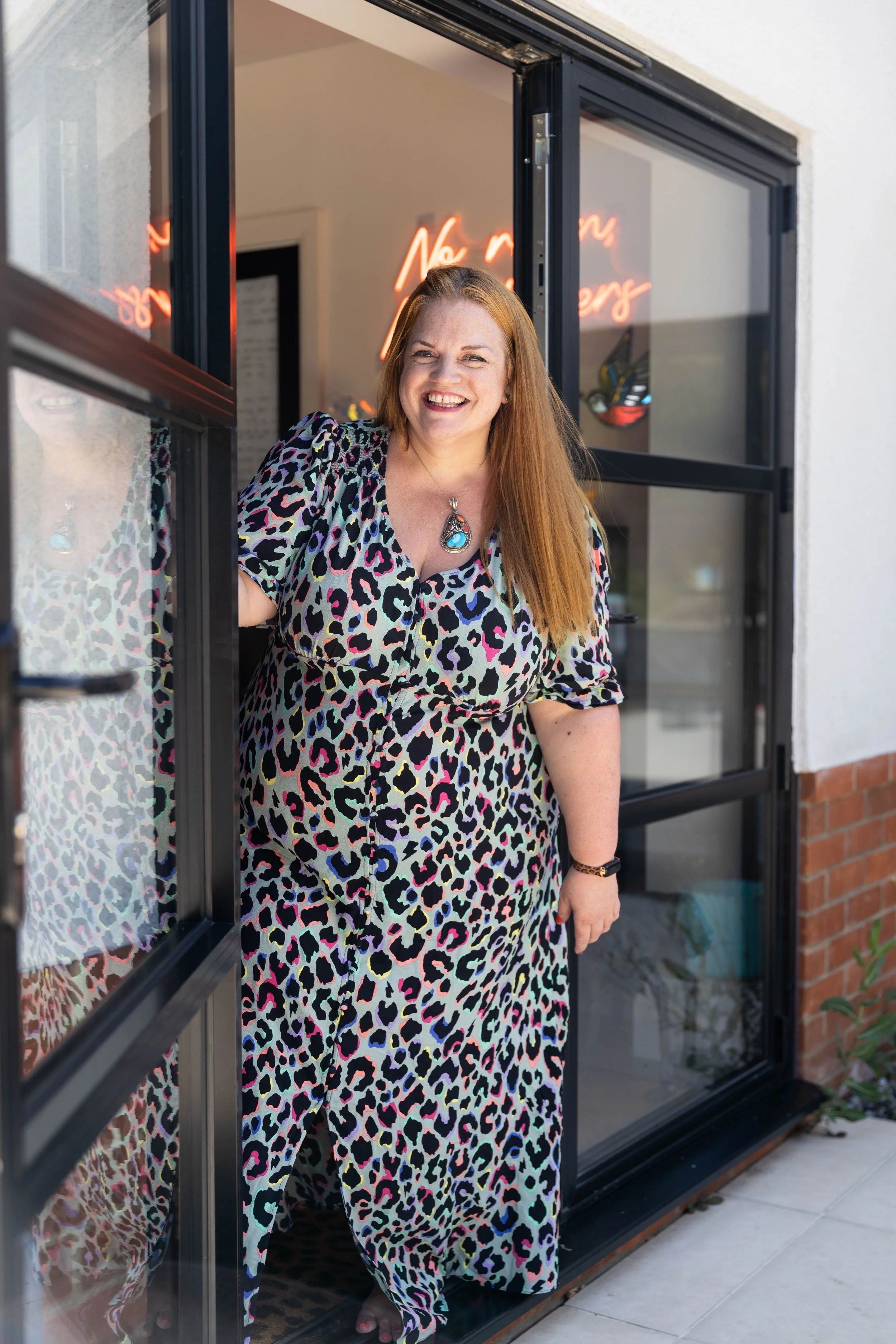 A woman with long red hair is standing in the doorway of a shop, smiling. She is wearing a colorful leopard print dress and a large turquoise necklace. The shop has a glass door with a neon sign inside that is partially visible.