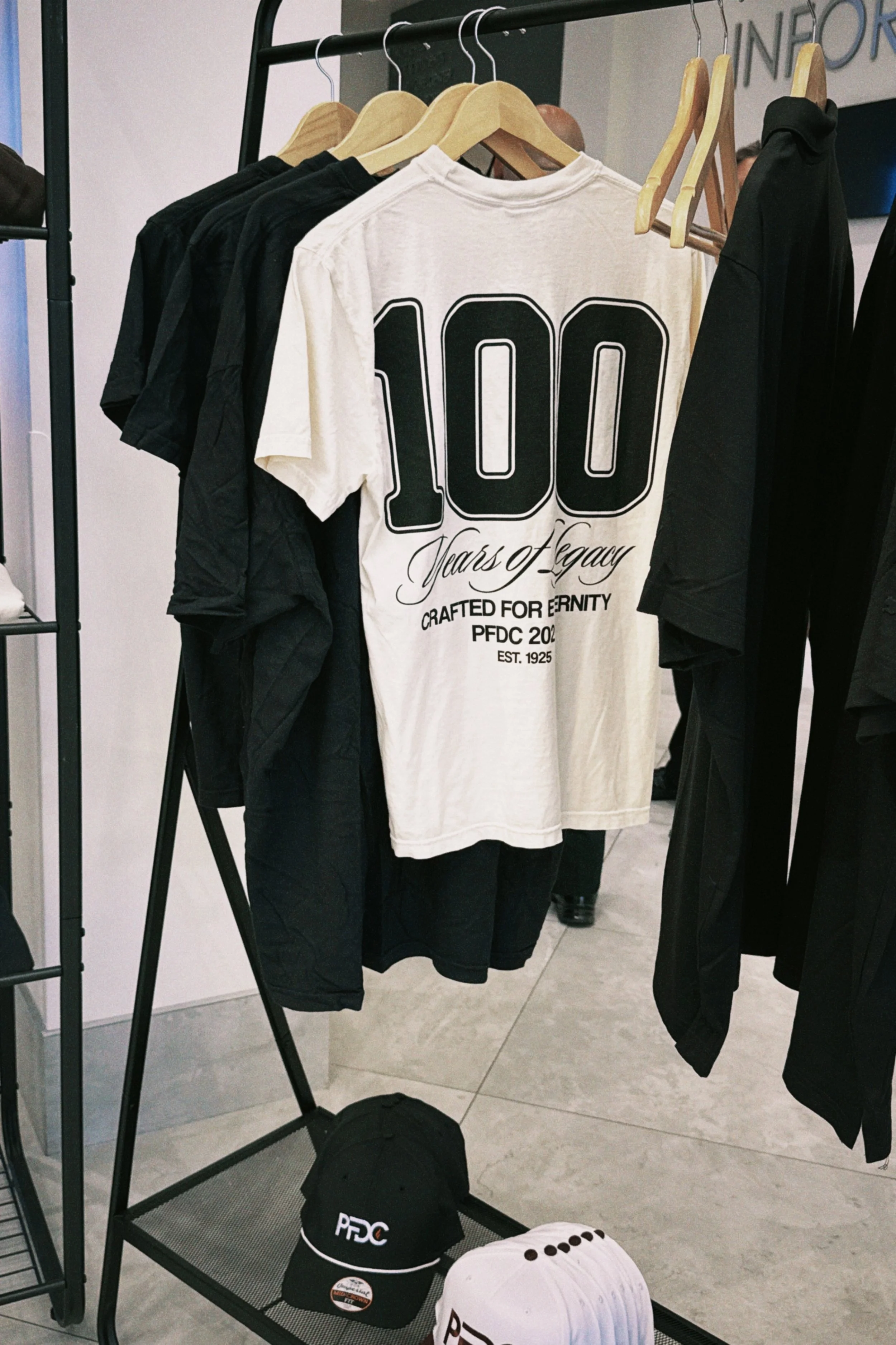 Clothing rack displaying black and white T-shirts, with a prominent white T-shirt celebrating 100 years of legacy and featuring the text and logo for PFDC; also includes black caps with the PFDC logo placed on the floor beneath the rack.