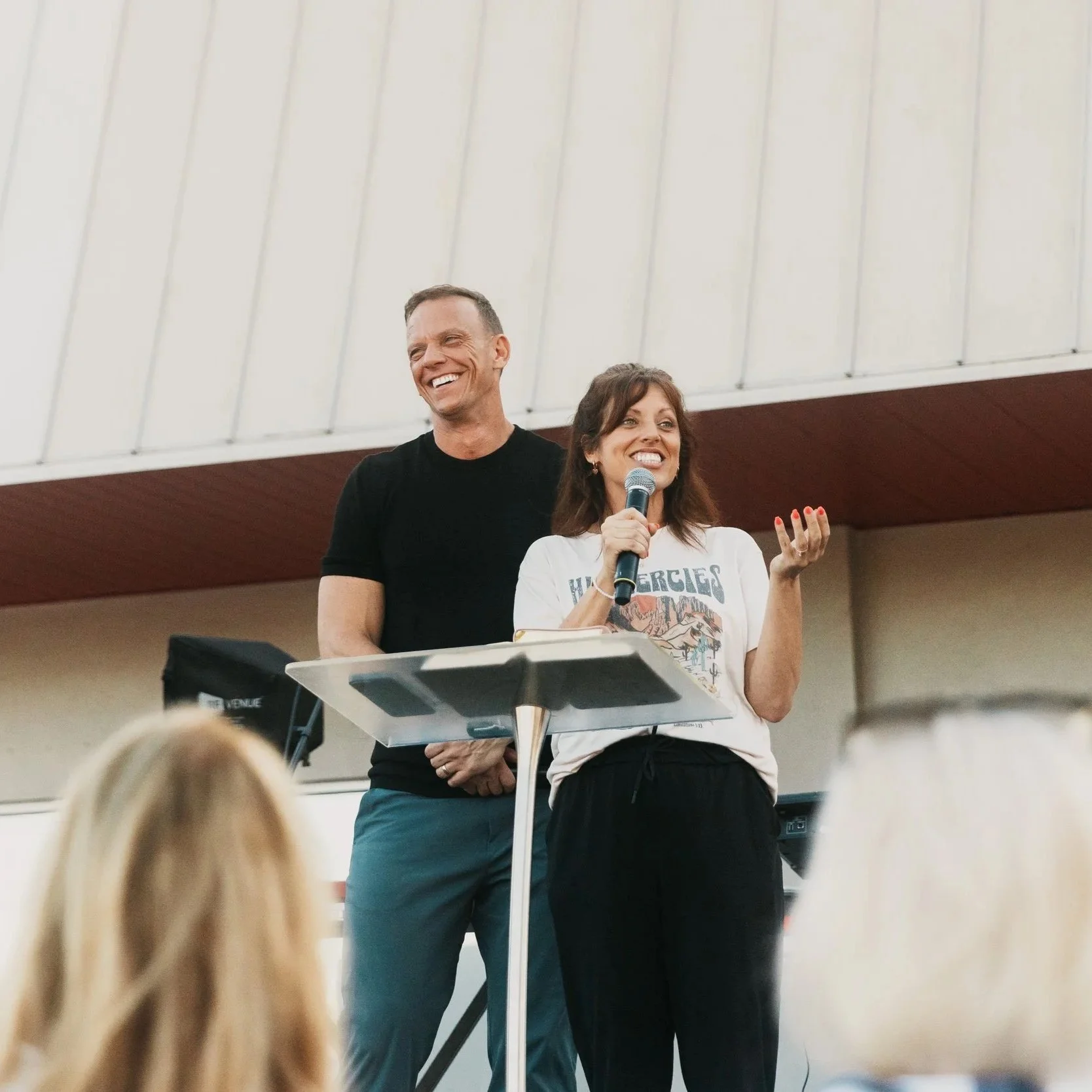 Two people standing at a podium, smiling and speaking at an indoor event, with a musician's keyboard visible in the foreground and audience heads in the bottom of the picture.