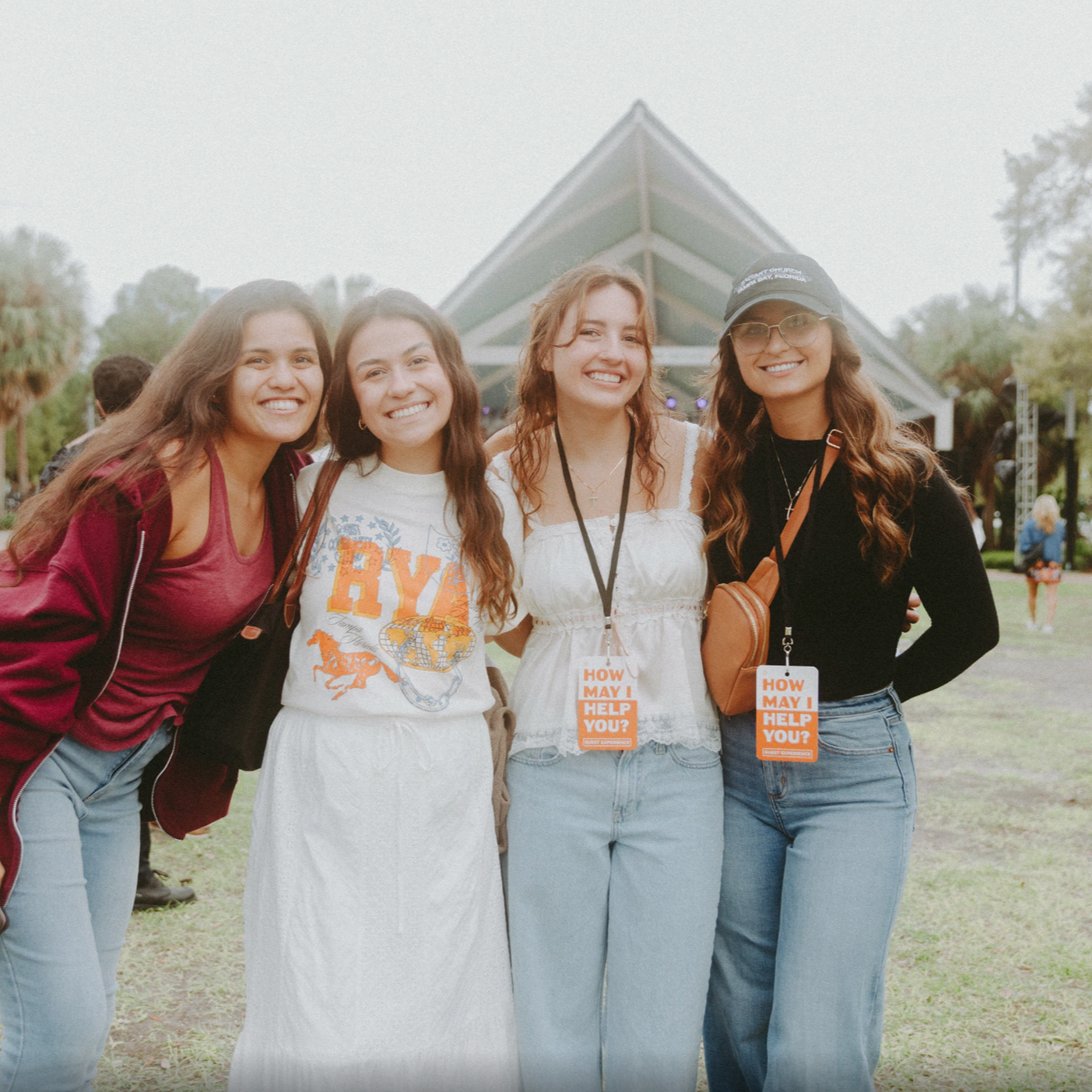 Four women standing together outdoors at an event, smiling, with a stage and trees in the background.