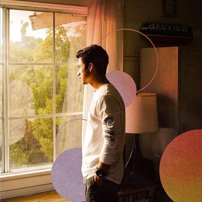 A young man stands in profile by a window, looking outside during sunset, with sunlight illuminating his face and white long-sleeve shirt in a cozy room.