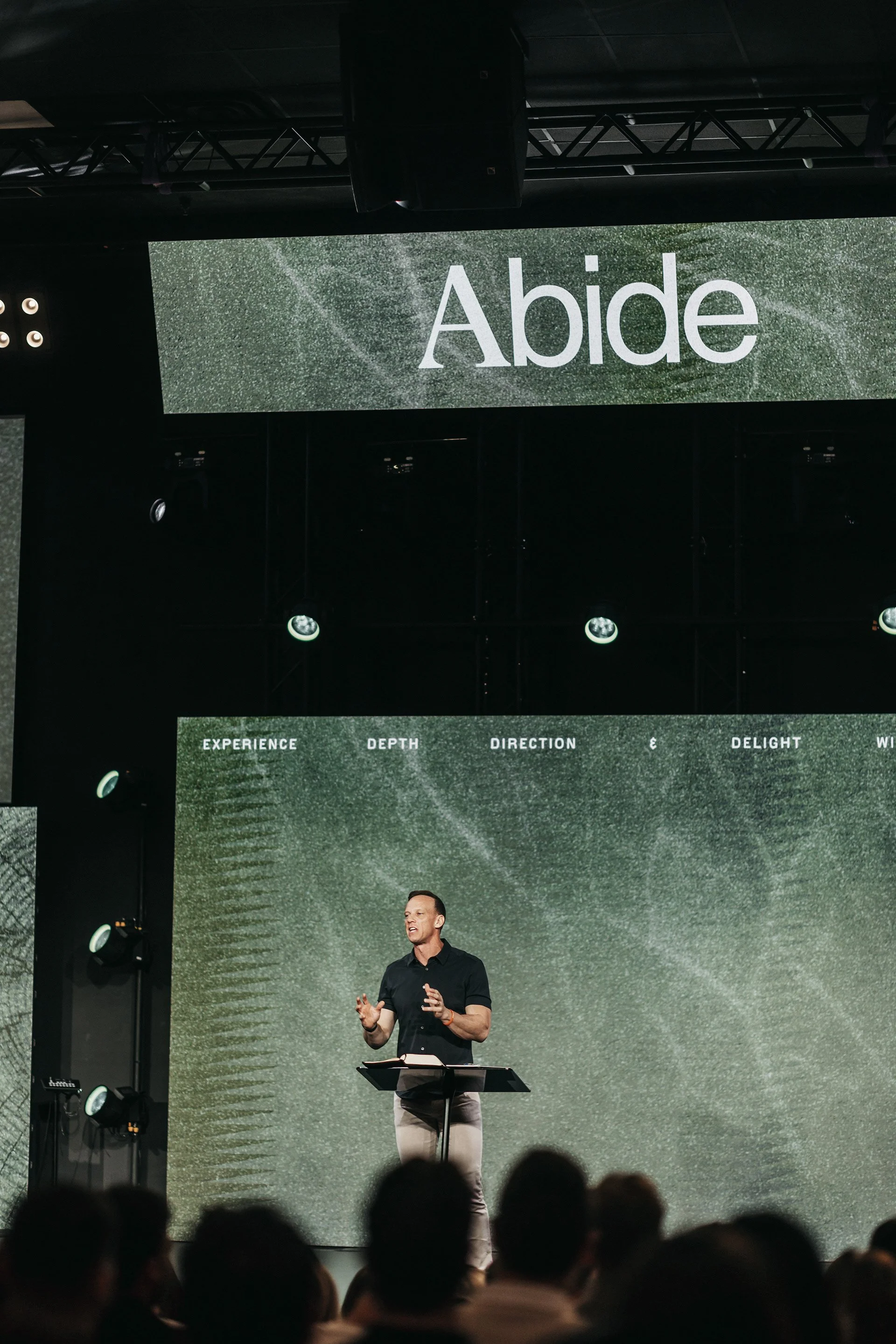 A man stands on stage in front of a large screen displaying the word 'Abide' and other words like 'Experience,' 'Depth,' 'Direction,' and 'Delight.' The audience is visible in the foreground.