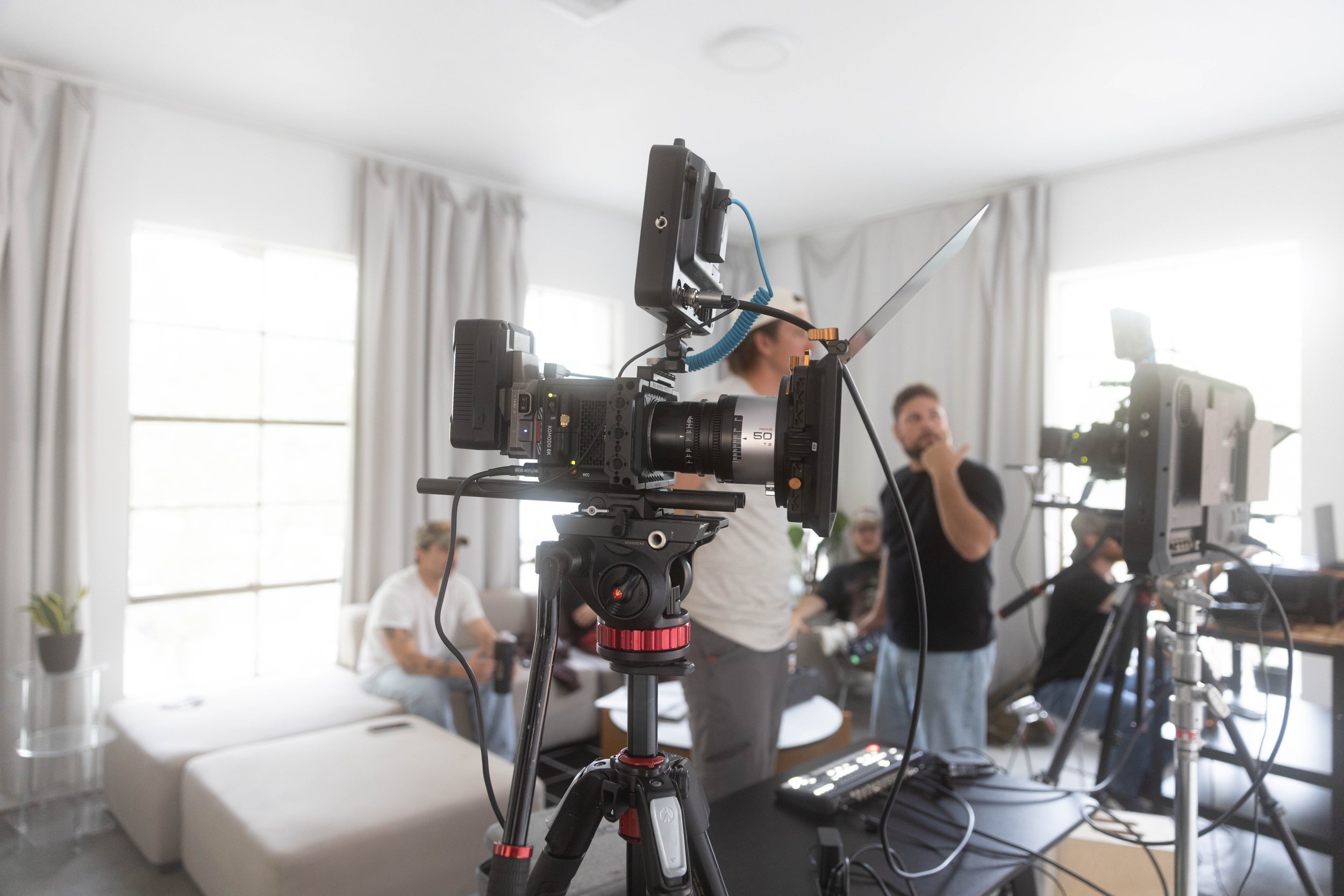 Camera on tripod in a bright room with large windows, people working at desks in the background, and a man standing and talking.