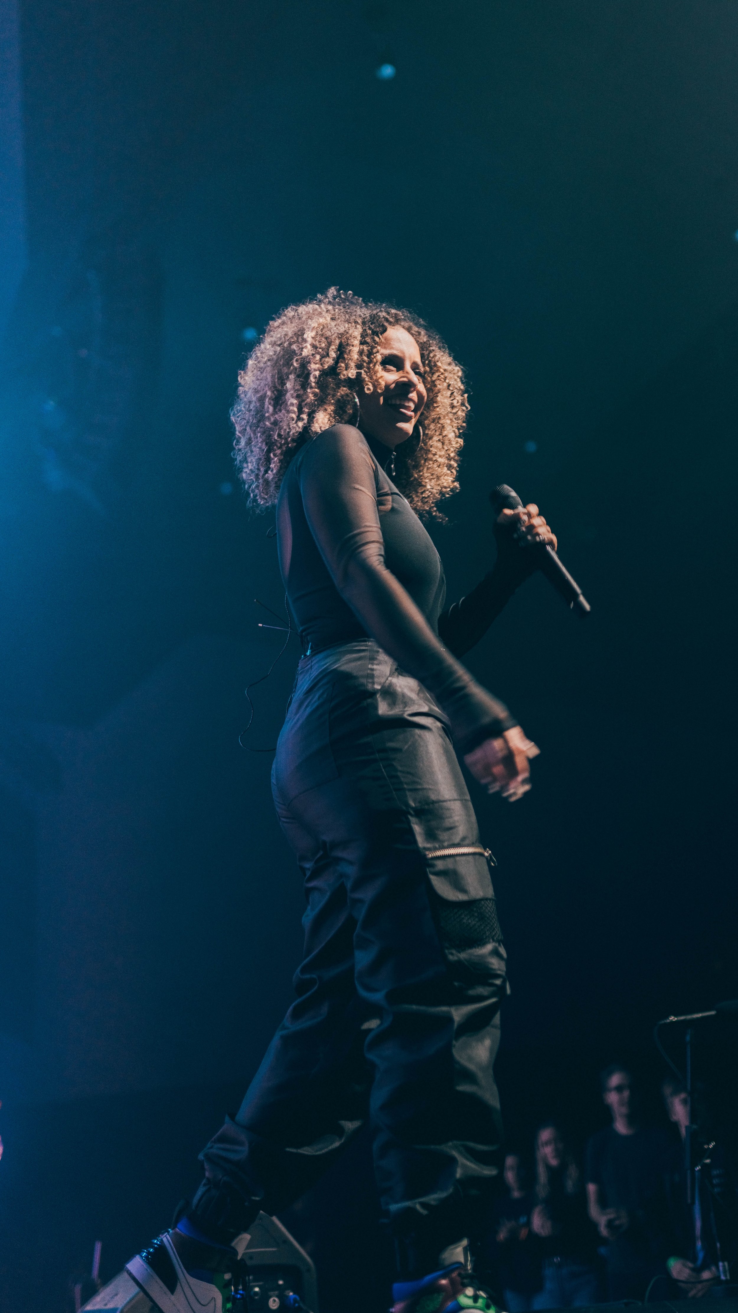 A woman with curly blonde hair singing into a microphone on stage, dressed in a black long sleeve top and black cargo pants, with audience members visible in the background.