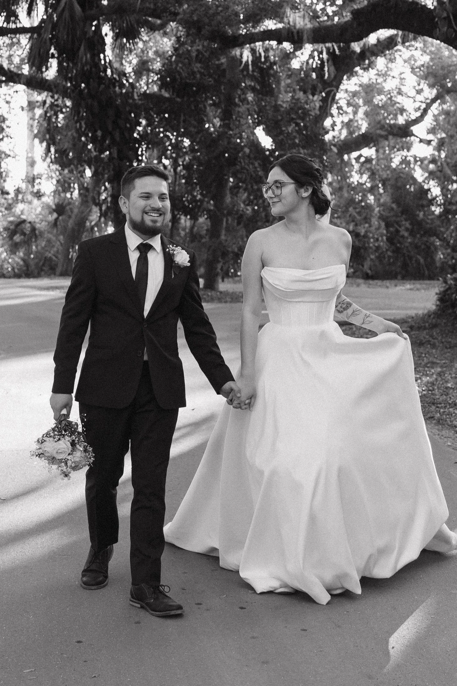 A black and white photo of a newlywed couple walking hand in hand outdoors, surrounded by trees. The groom is smiling and wearing a dark suit with a boutonniere, holding a bouquet of flowers. The bride is wearing a strapless wedding gown and glasses, looking at the groom with a smile.