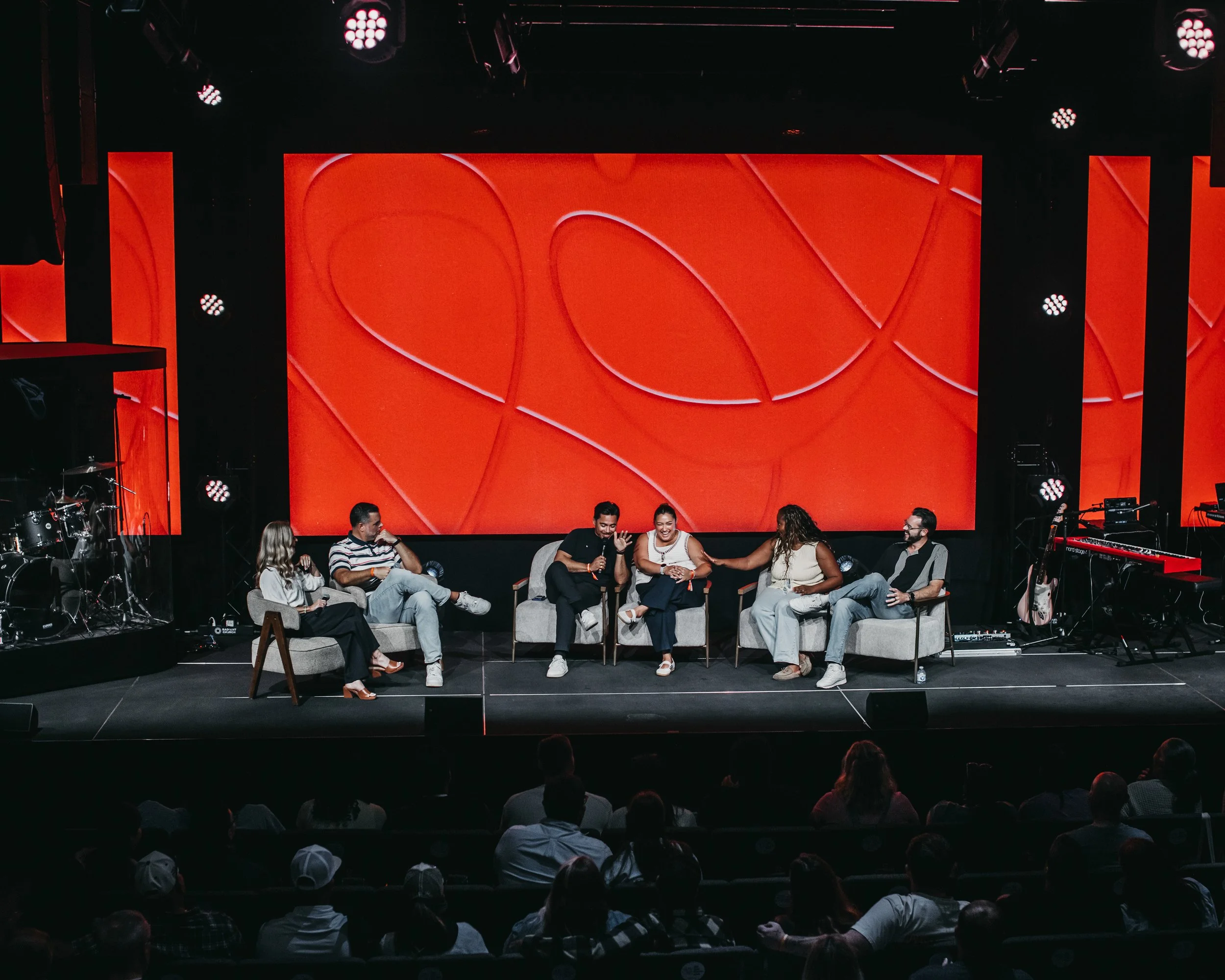 People sitting on chairs on stage during an event with a large red abstract background on a screen behind them, musical instruments on the side, and audience members in the foreground.