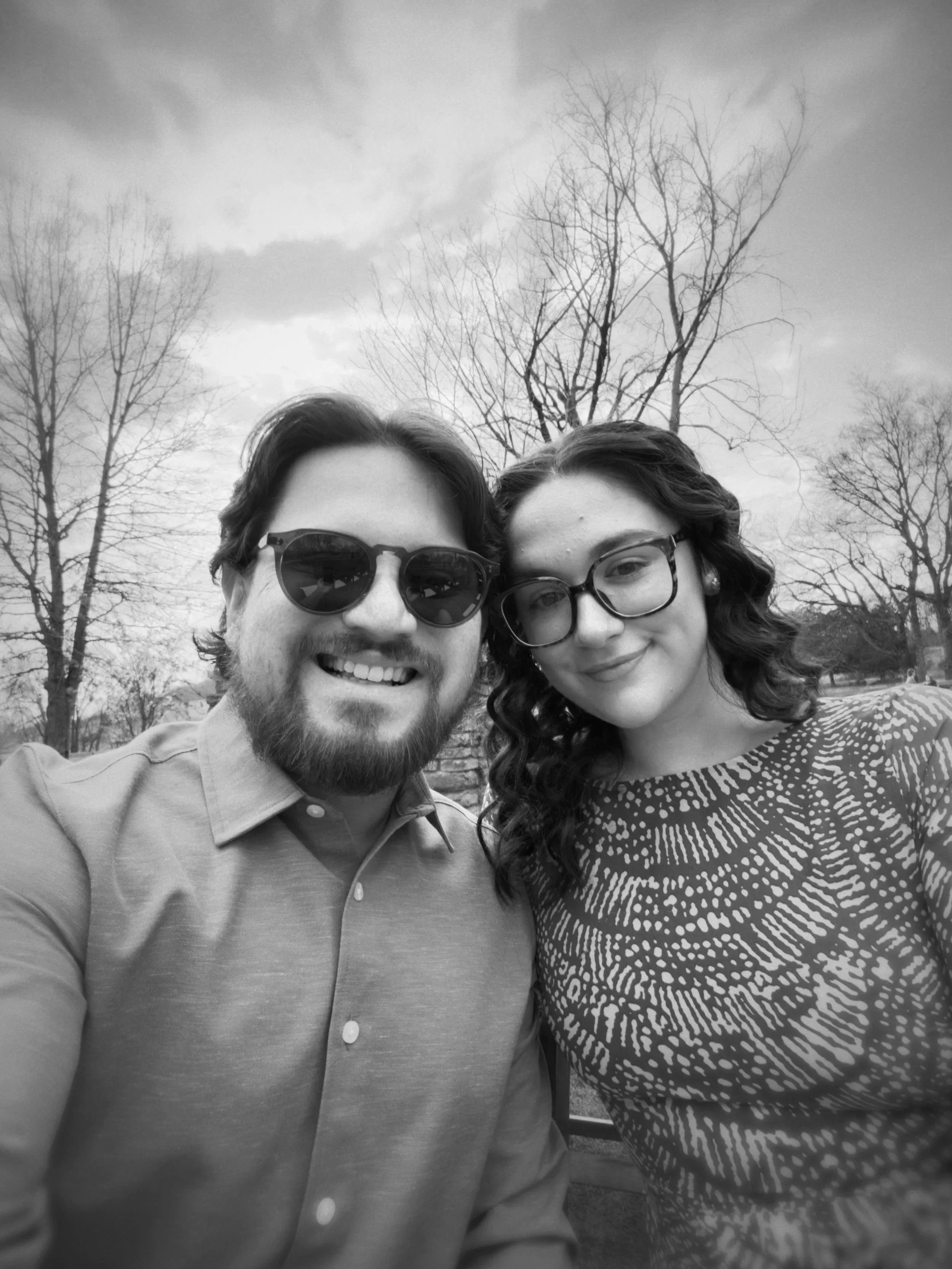 A black and white photo of a man and a woman taking a selfie outdoors. The man is wearing sunglasses and a collared shirt, and the woman has glasses and wavy hair. Behind them are leafless trees and a cloudy sky.