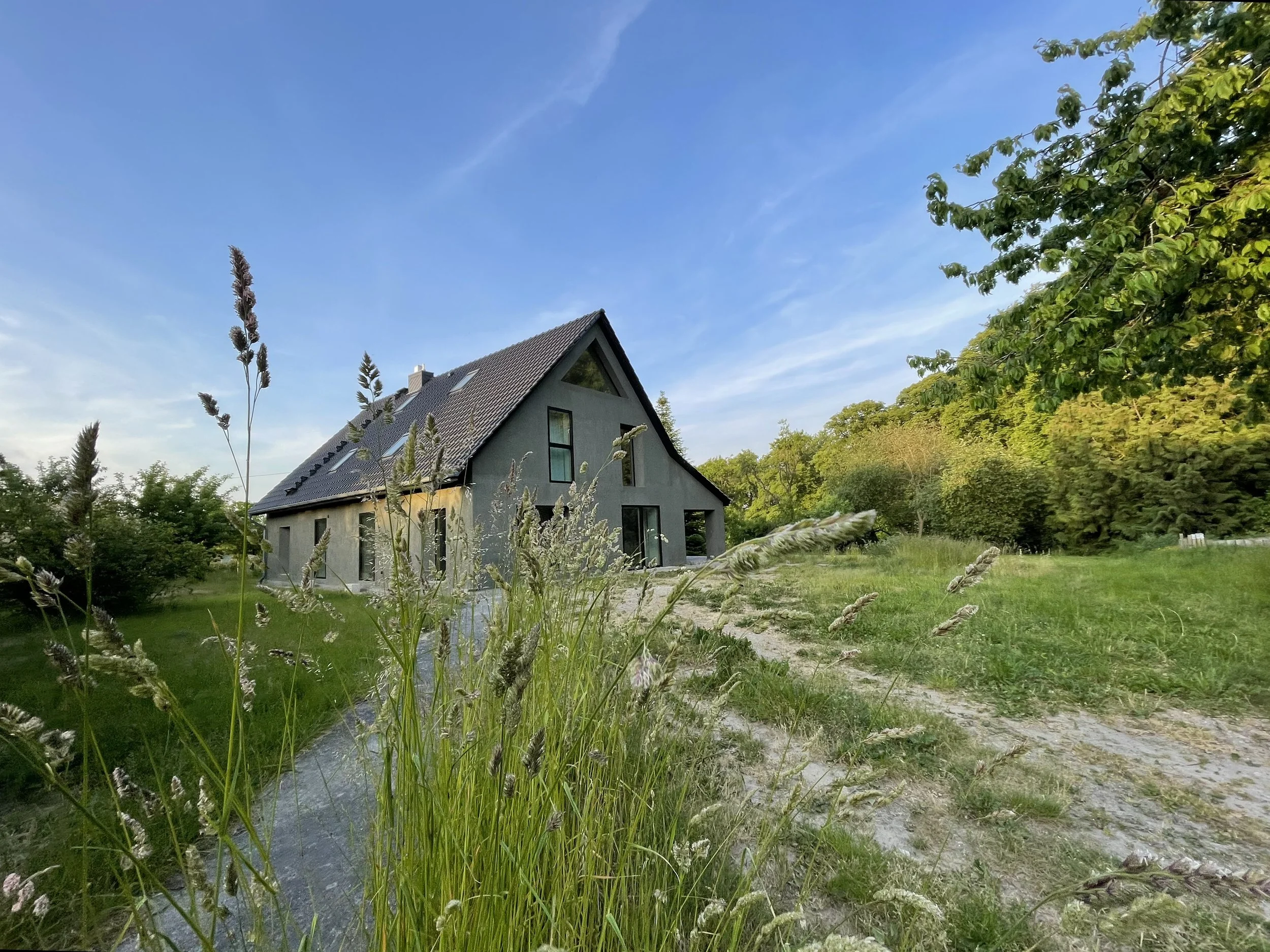 Mellenau 9. A modern house with a steep black roof and large windows, surrounded by green trees and grass, under a blue sky with some clouds.