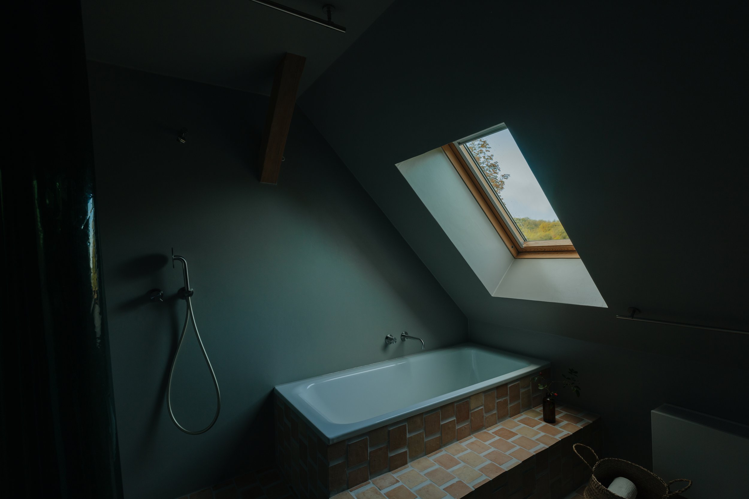 A bathroom with a bathtub beneath a skylight window, dark walls, and a brick platform foundation, with a showerhead on the wall.
