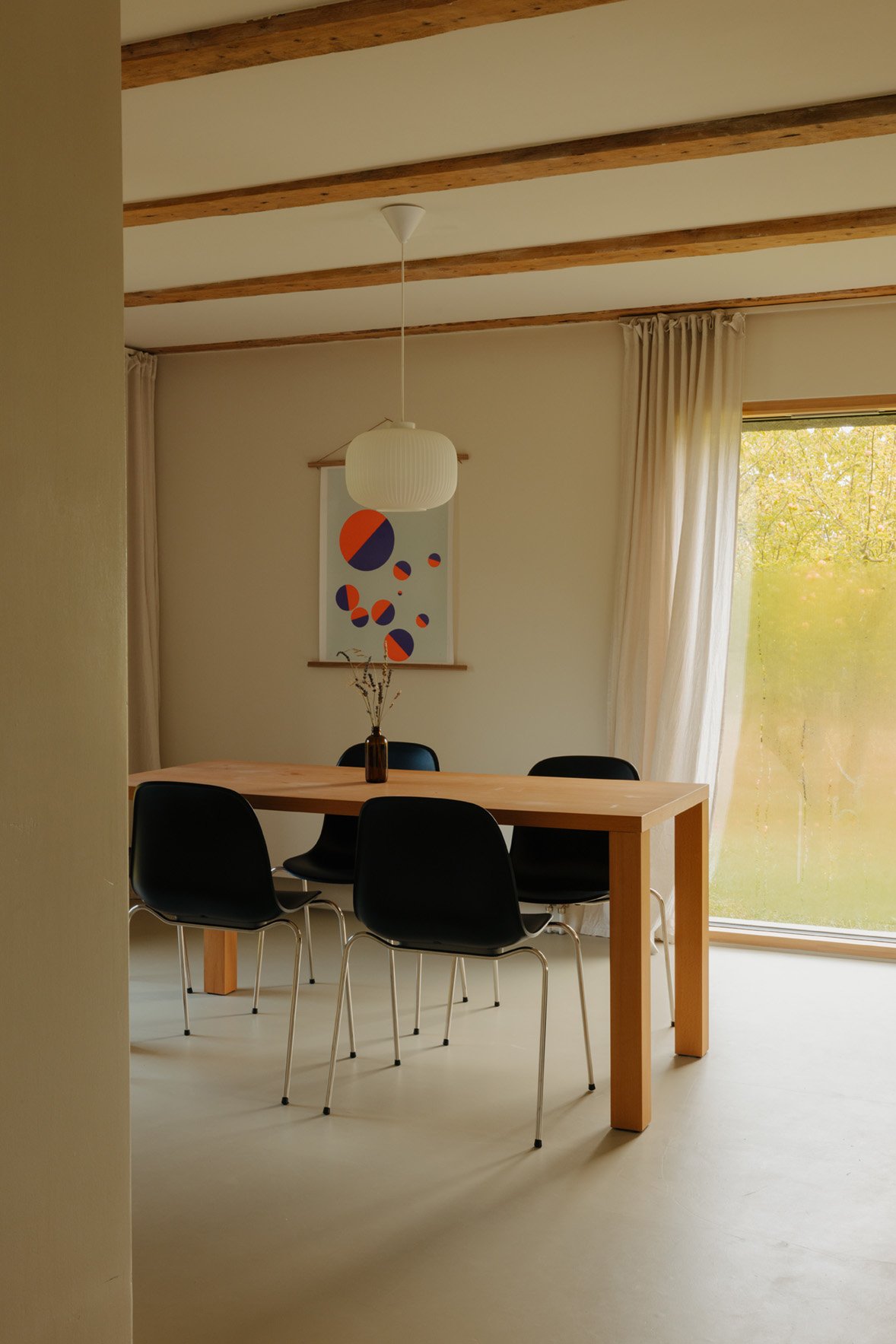 A dining room with a wooden table, four black chairs, a white paper lantern ceiling light, beige curtains, a large window showing trees outside, and modern artwork on the wall.