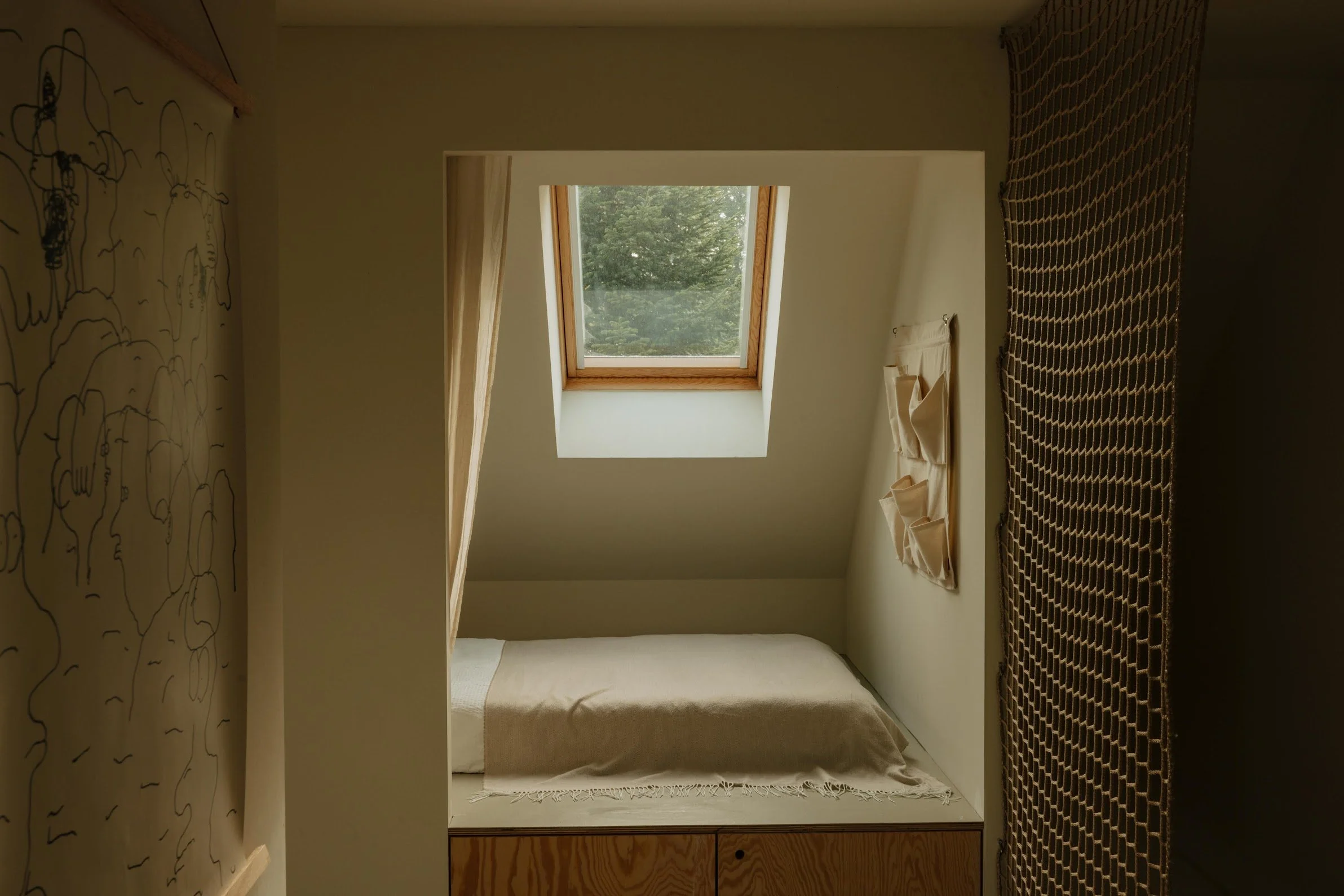 Small cozy bedroom with a bed underneath a skylight window, natural light coming through, beige bedding, and decorative wall hangings.