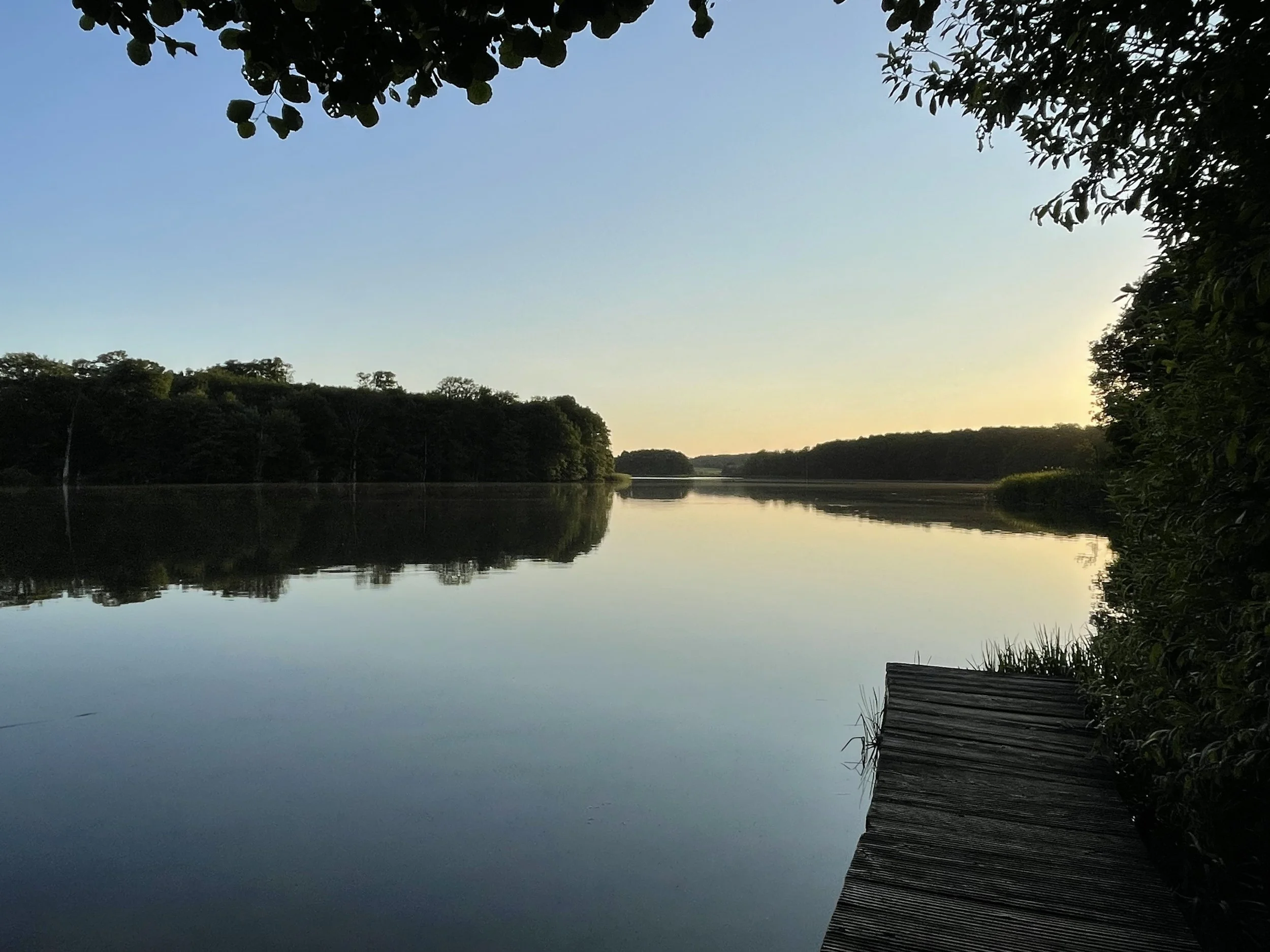 A serene lake scene during sunset at sunset with calm water reflecting the blue sky and surrounding trees, and a wooden jetty extending into the water on the right side.