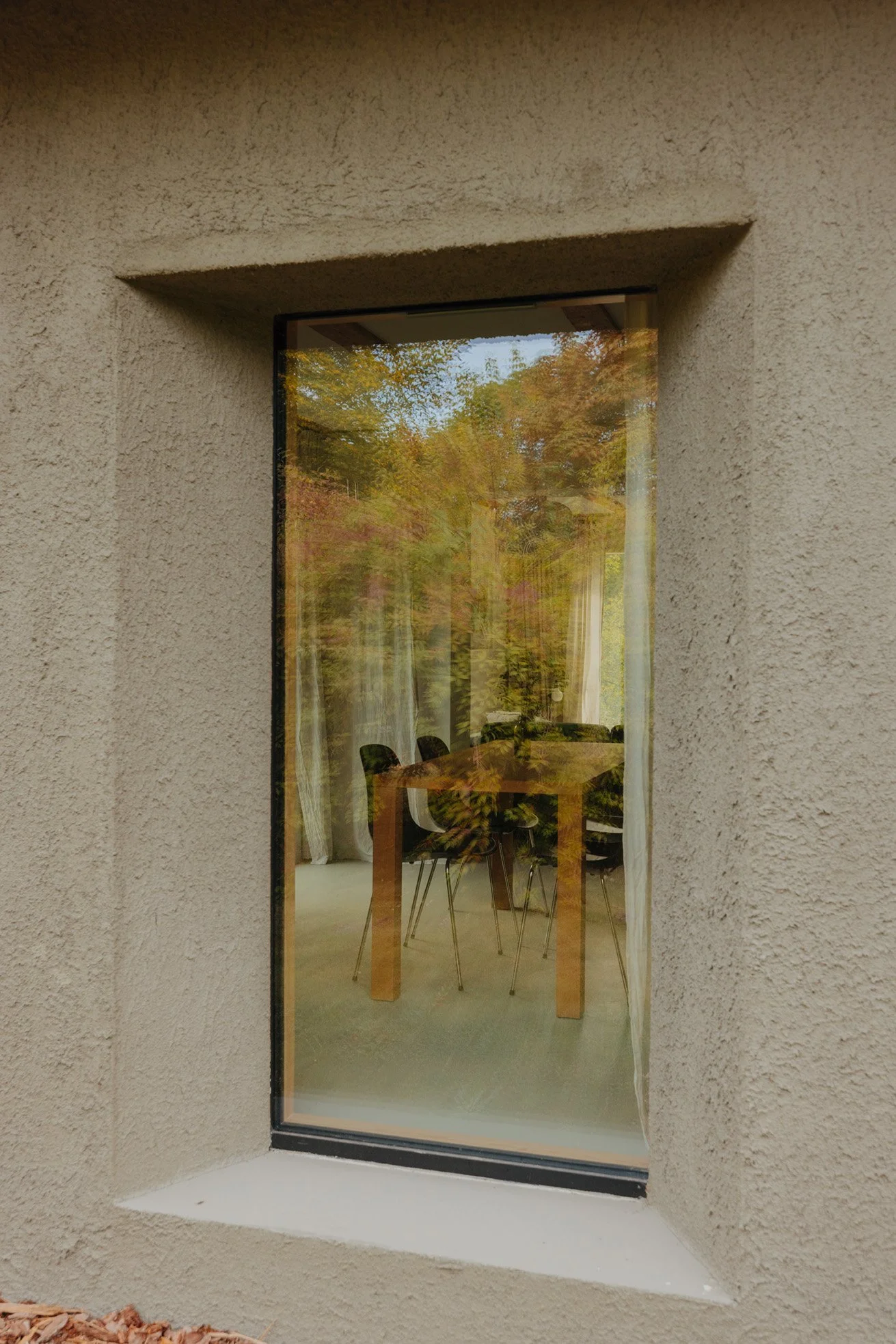Mellenau 9. View through a window showing a dining room with a wooden table, six black chairs, and trees with autumn-colored leaves outside.