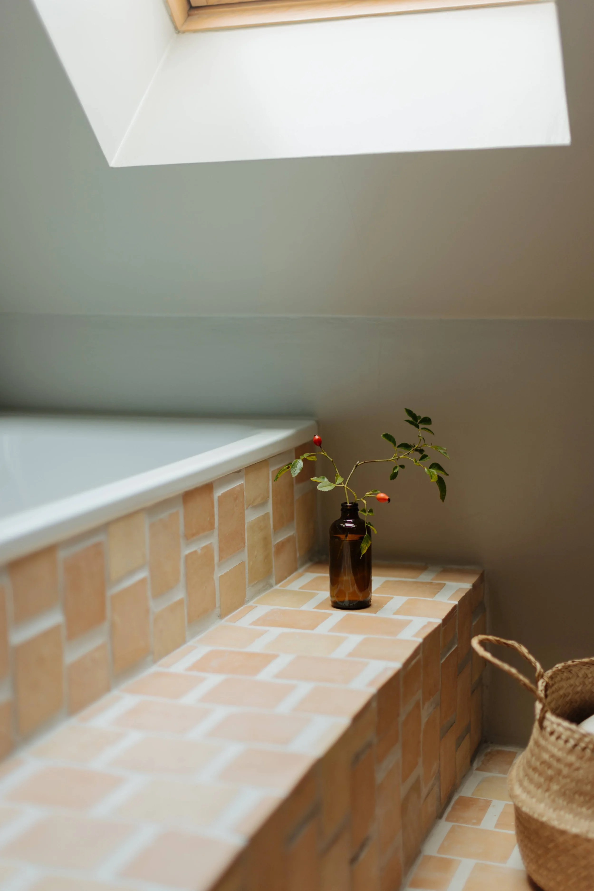A brown glass bottle with green leaves and red berries, placed on a brick ledge in front of a bathtub.