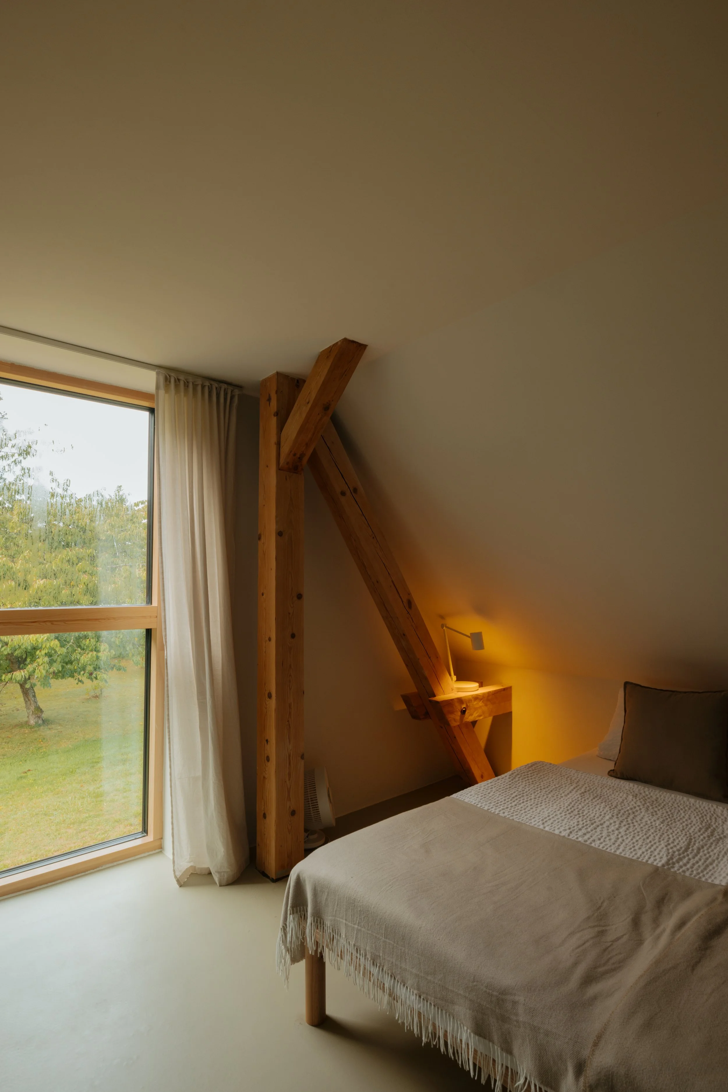 A bedroom with a bed, beige blanket, and pillow. Wooden beams and a small lamp on a side table are near the bed. Large window with white curtains shows greenery outside.