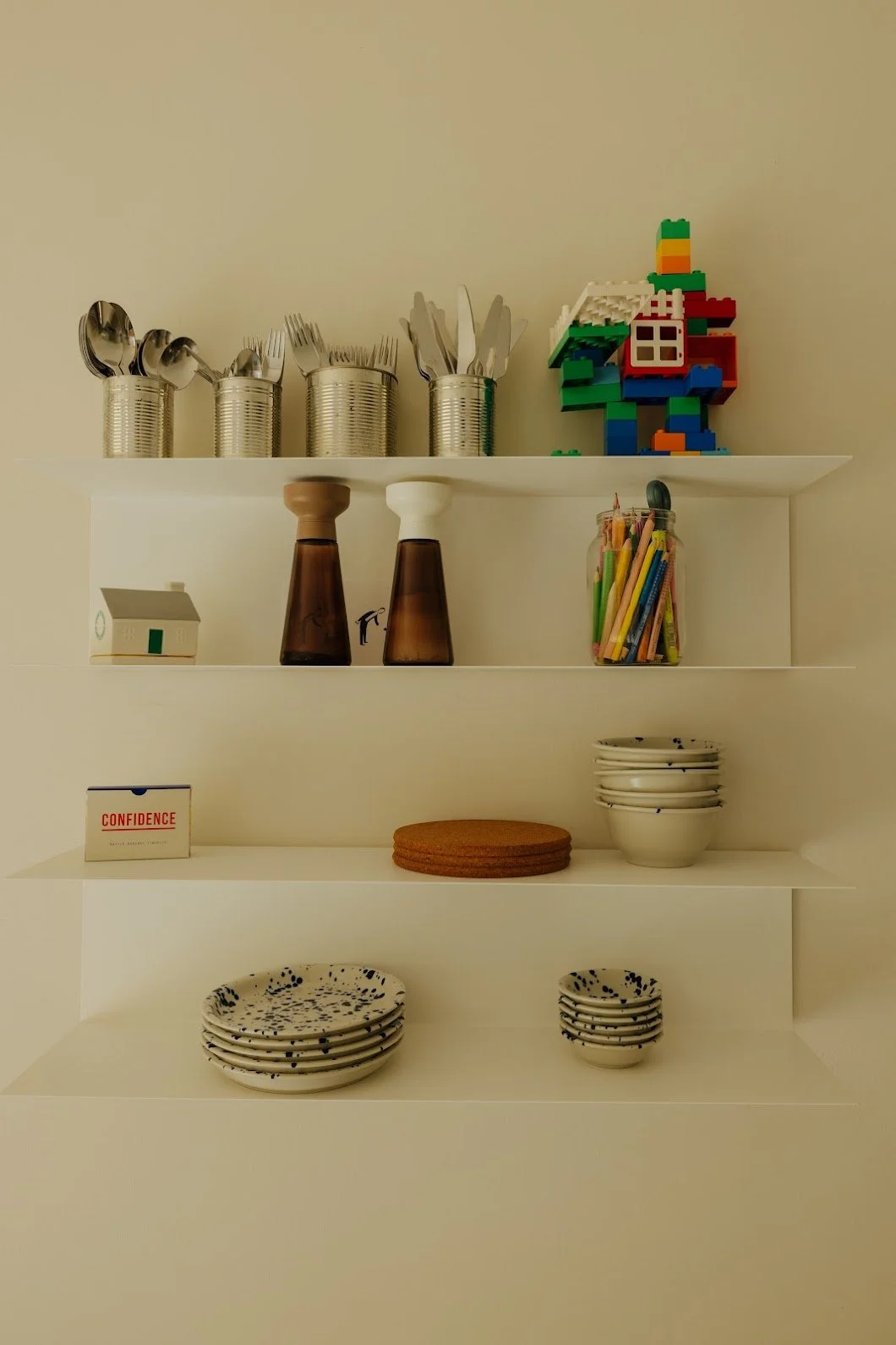 Mellenau 9. White wall-mounted shelves with kitchen items including silver cans with utensils, a LEGO house, condiment containers, a glass jar of colored pens, a small house figurine, stacks of patterned plates and bowls, and cork coasters.