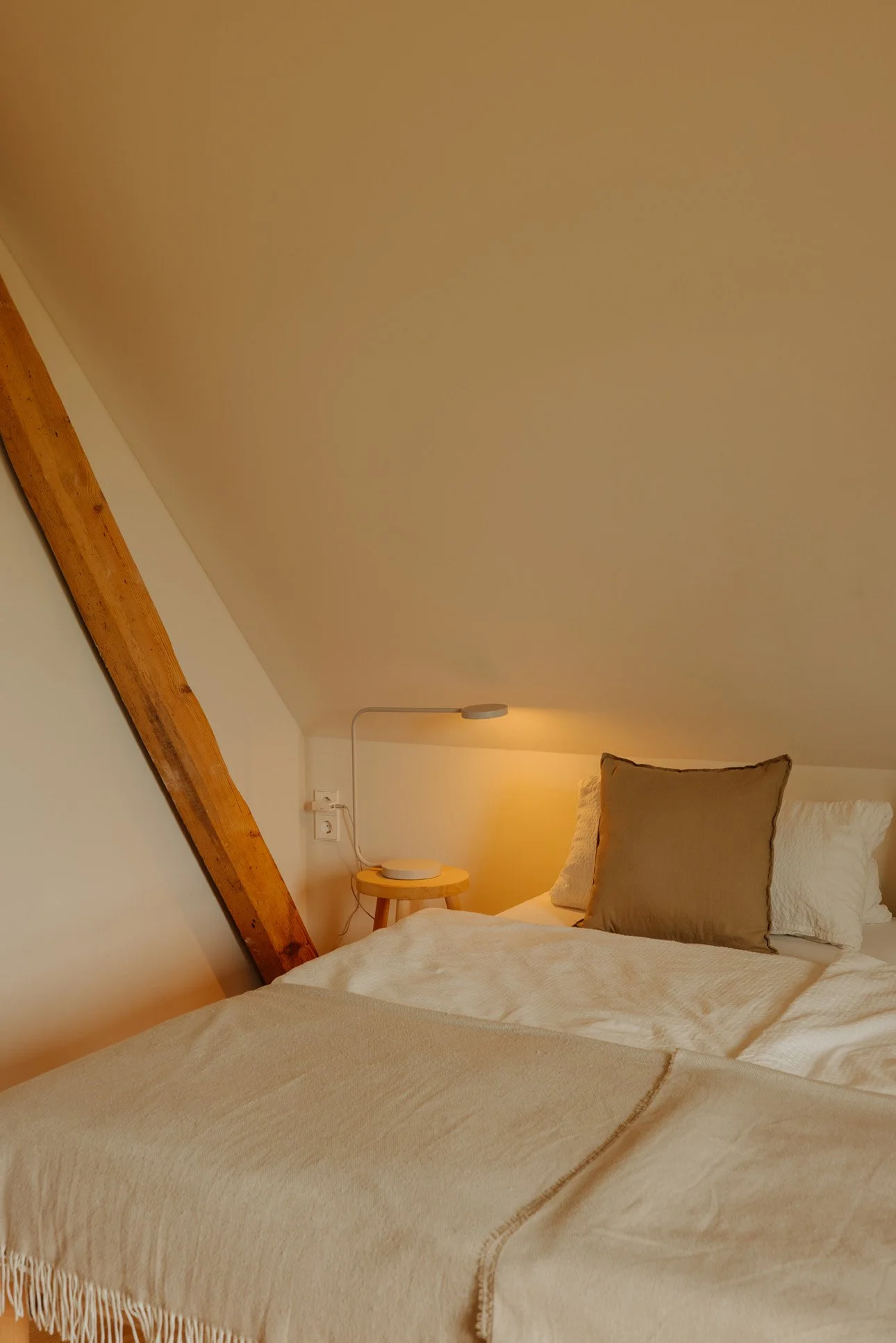 A cozy bedroom with a sloped ceiling, a bed with beige bedding and pillows, a small bedside table with a modern lamp, and a wooden beam on the wall.