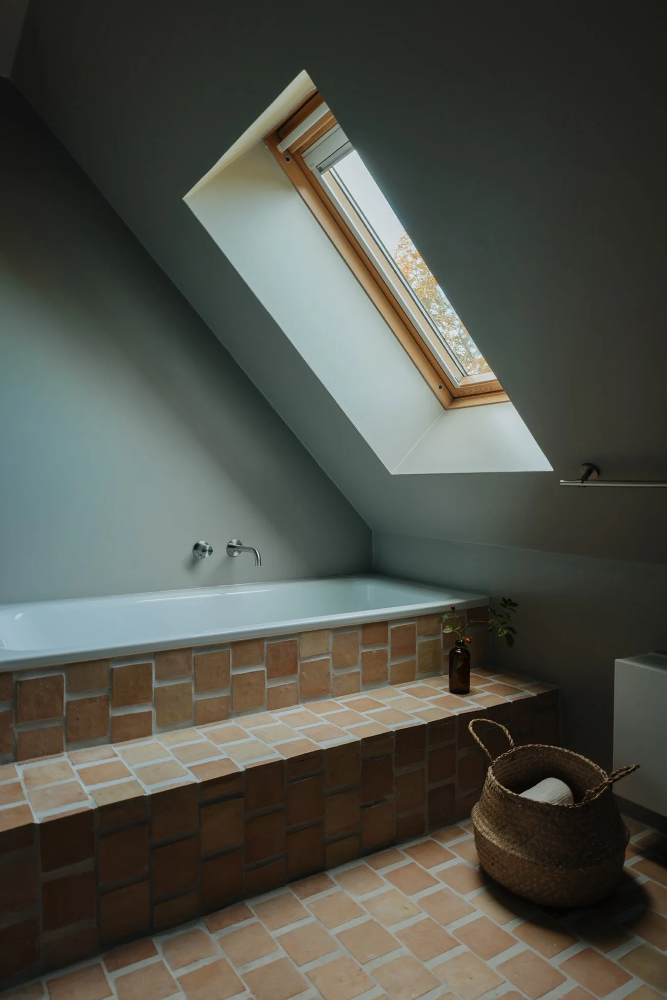 A bathroom with a skylight window in the ceiling, a bathtub with brick tile surround, a wicker basket with a towel inside, and a small plant in a dark bottle on the ledge.