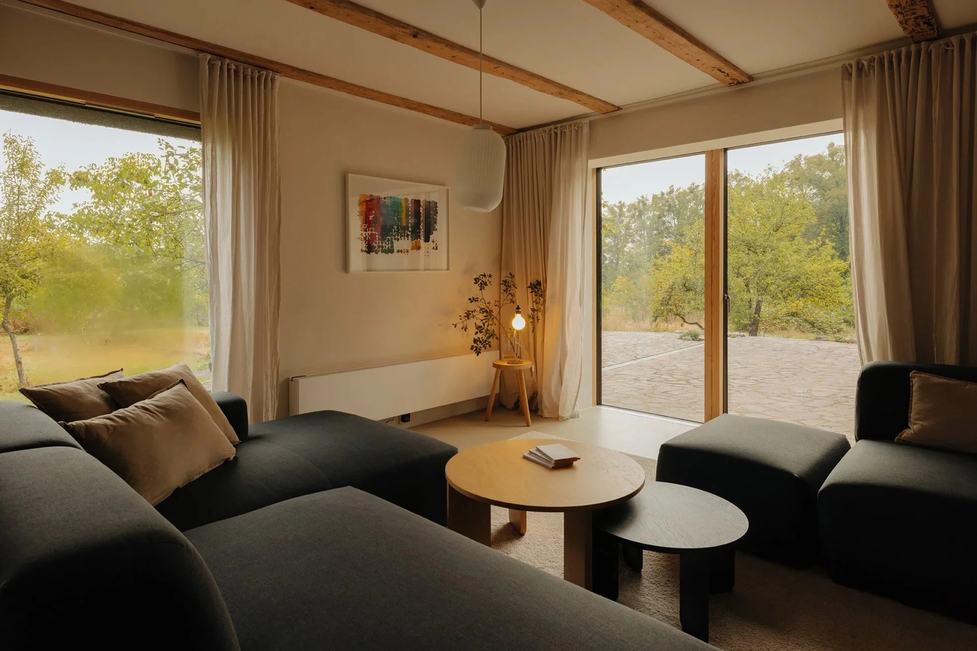 Living room with a large sofa, armchair, round coffee tables, large windows with curtains, view of trees outside, and a small side table with a lamp