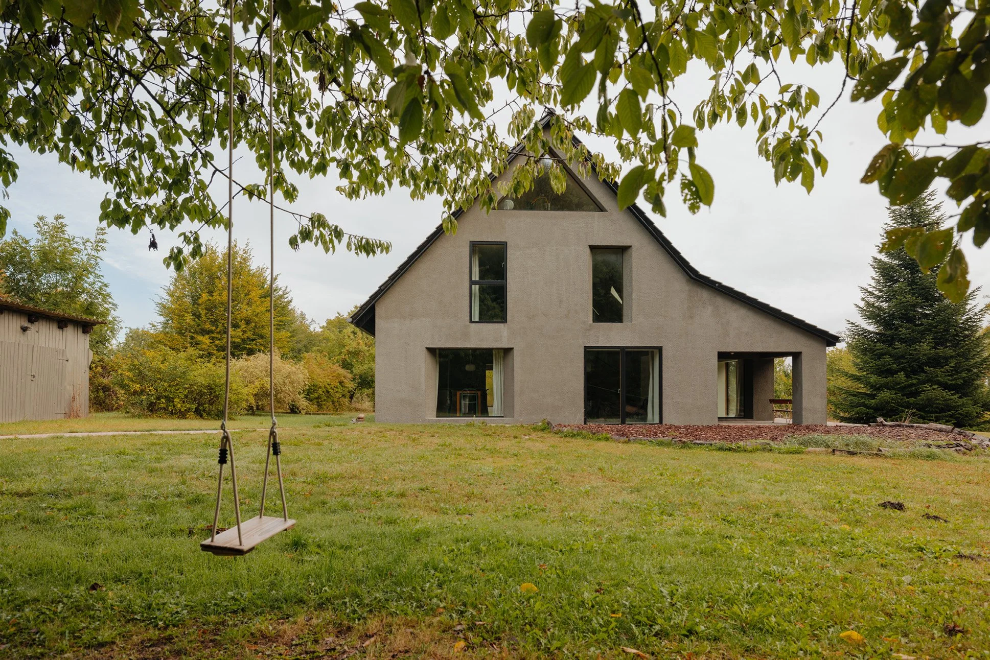 Mellenau 9. A modern house with large windows, surrounded by trees and a grassy yard, with a wooden swing hanging from a tree branch in the foreground.