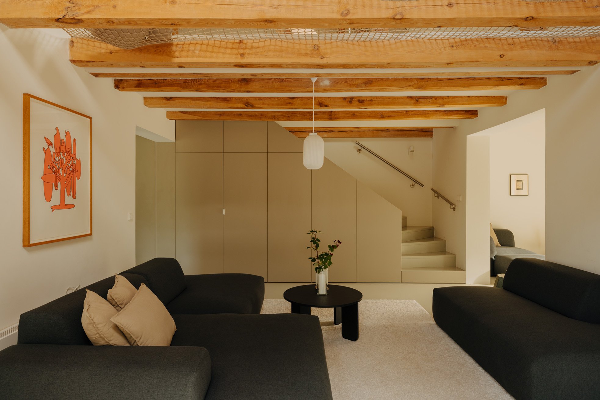 Living room with two black sofas, a round black coffee table with a white vase of flowers, wooden ceiling beams, a staircase in the background, a pendant light, and a piece of orange abstract art on the wall.