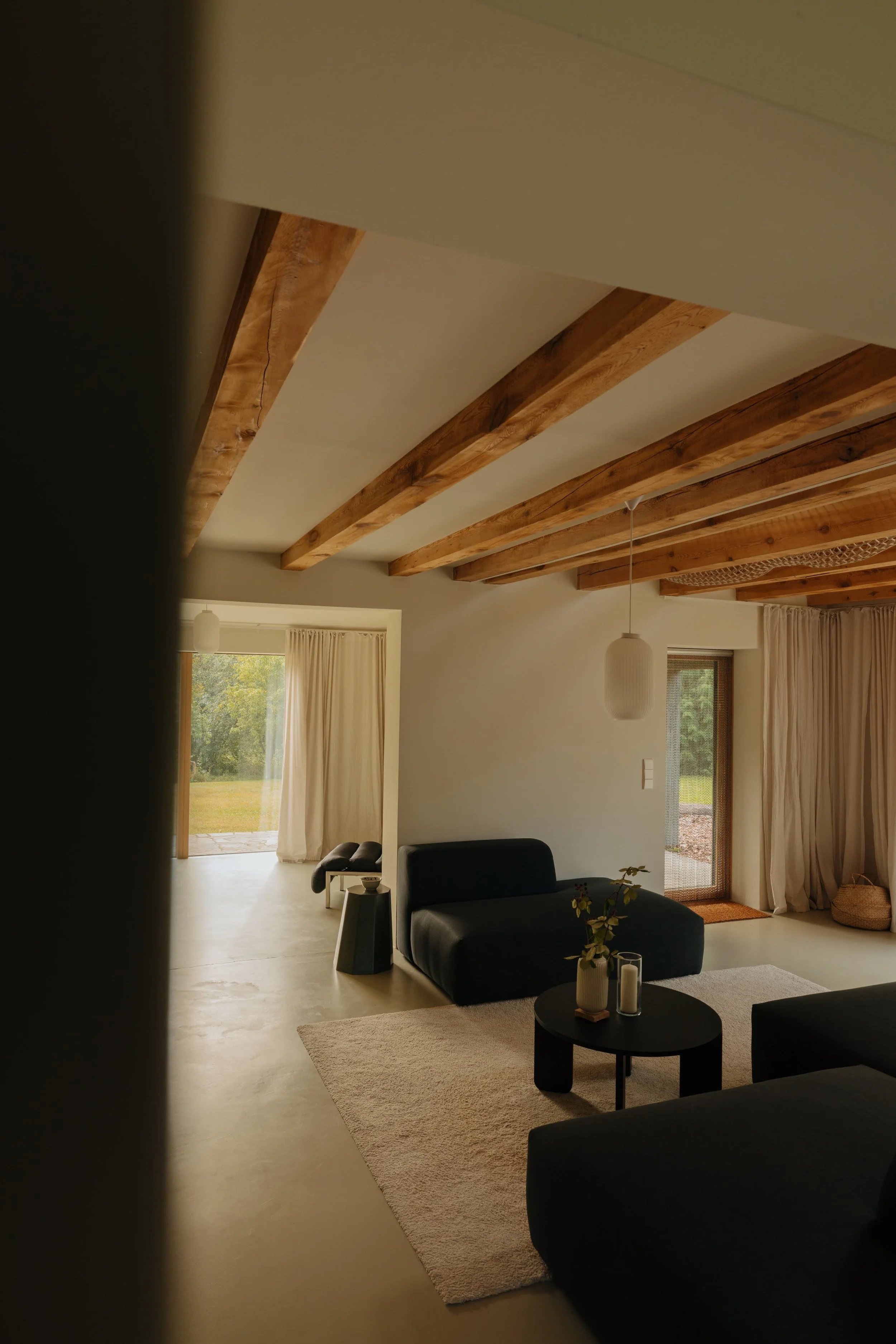 Living room with beige walls, wooden ceiling beams, black furniture, a round black coffee table with vases, and large windows with curtains opening to a green outdoor space.