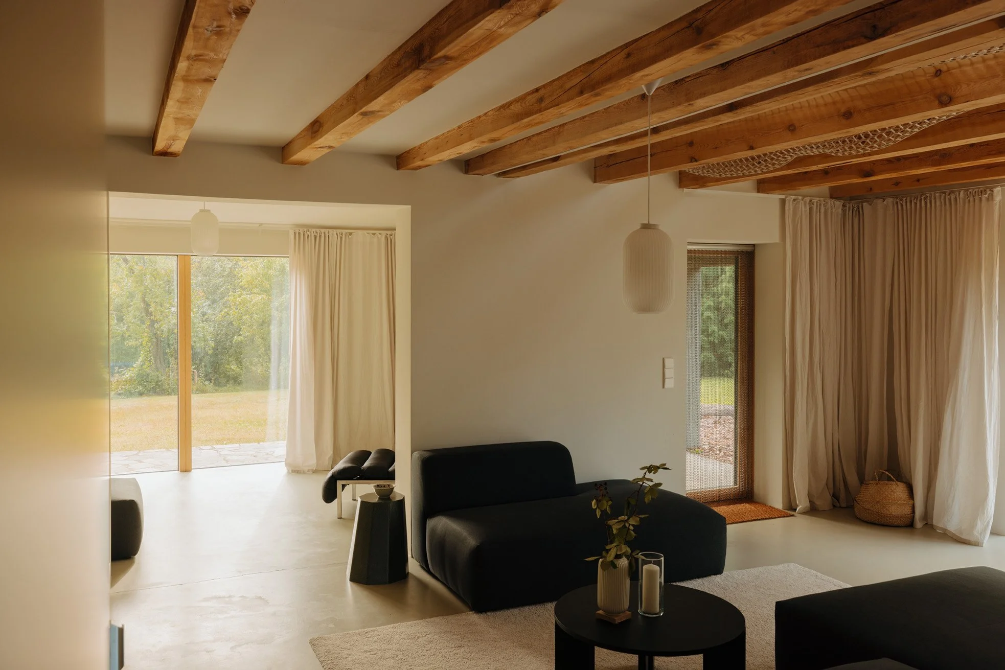 Living room with wooden ceiling beams, beige curtains, black sofas, a round black coffee table with candles and a plant, and large windows showing a grassy outdoor area.