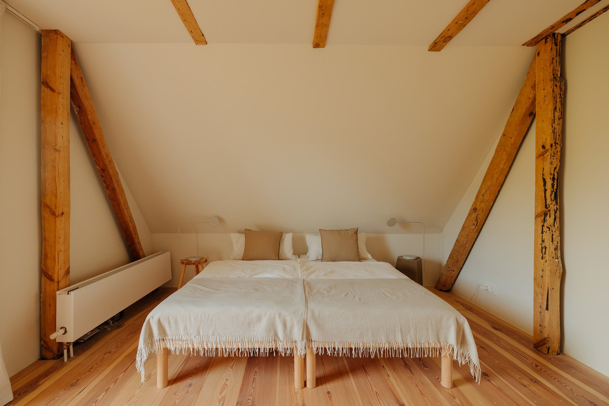 A minimalist bedroom with a double bed, neutral-colored pillows, and throw, set against a white wall with exposed wooden beams in the ceiling and walls.