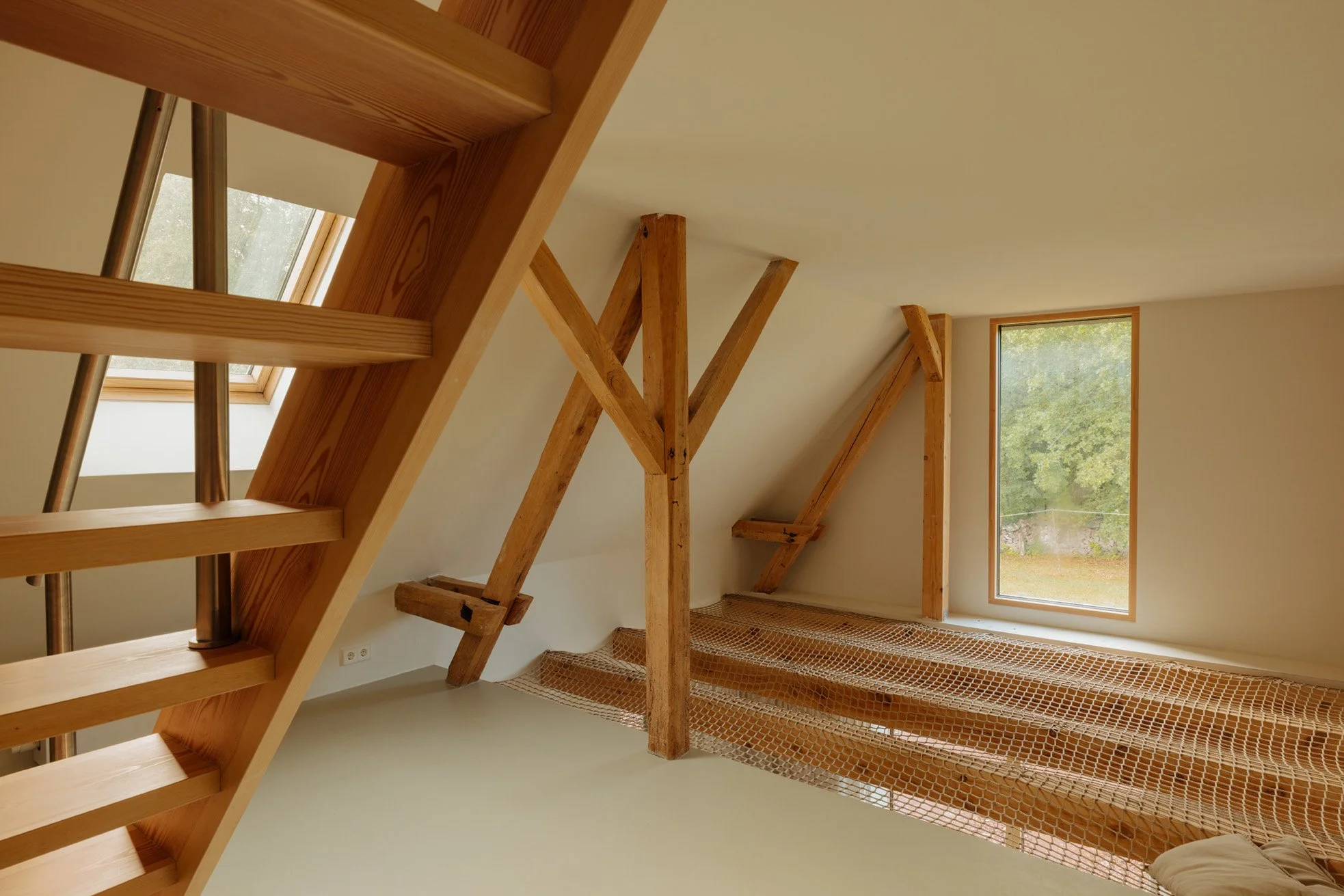 View of an attic room with wooden beams and a staircase, large windows showing trees outside, and a protective netting on the floor.