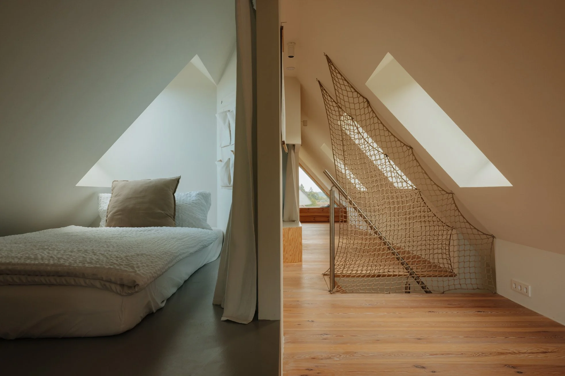Split view of an attic room: on the left, a cozy sleeping space with a bed under a triangular skylight, beige pillows, and white bedding; on the right, an open area with a wooden floor, triangular skylight, a safety net near an open staircase, and a window showing a landscape outside.