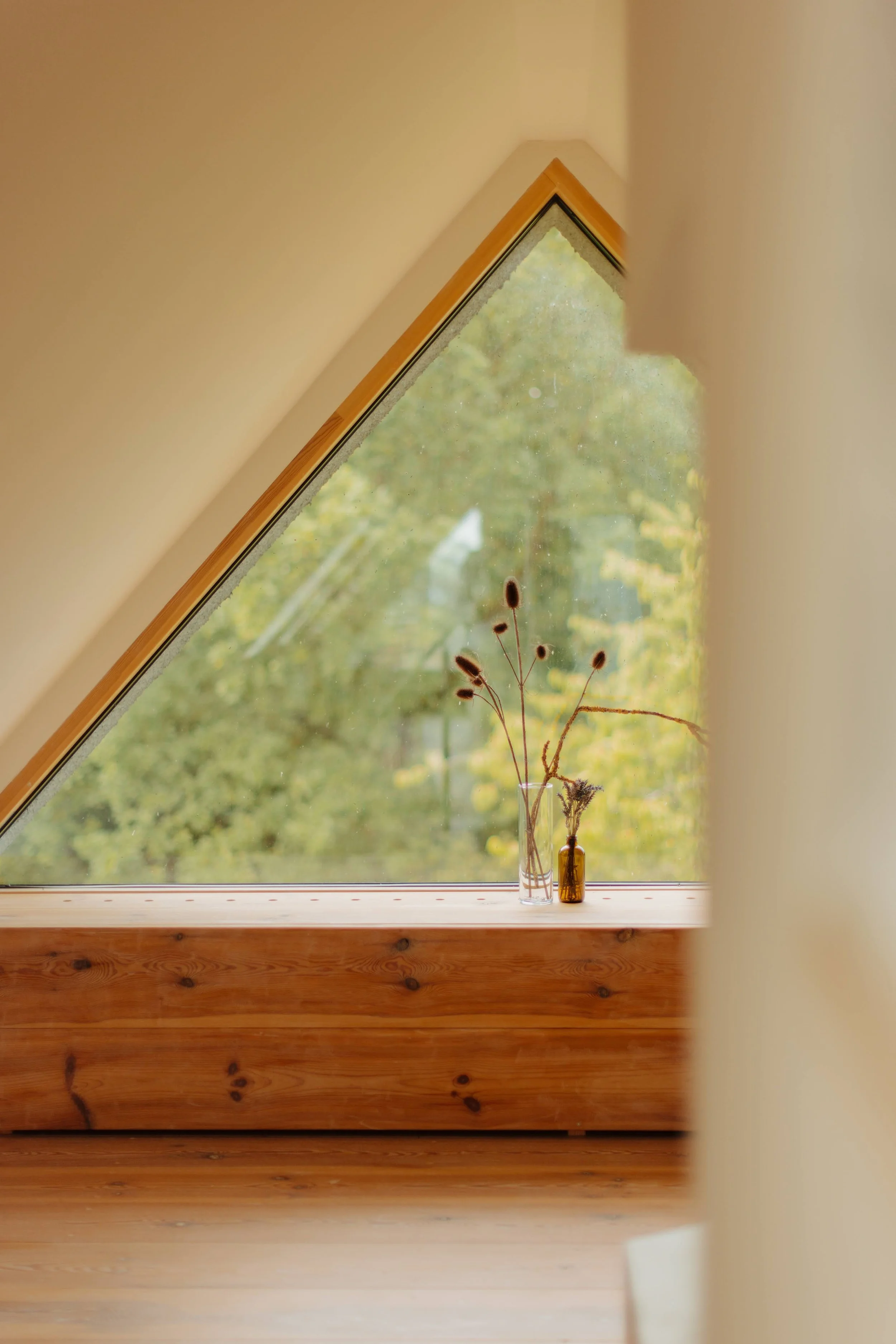 View of a skylight window with dried flower arrangements in small bottles on the windowsill, outside a forest is visible through the glass.