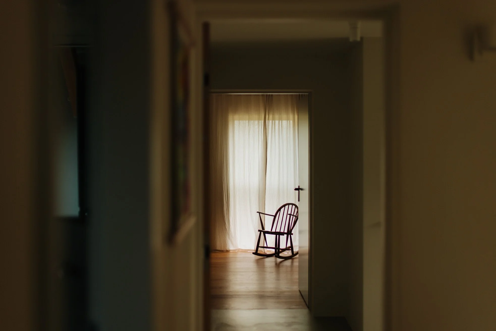 A purple rocking chair in front of cream curtains covering a window, as seen through a doorway in a dimly lit corridor.