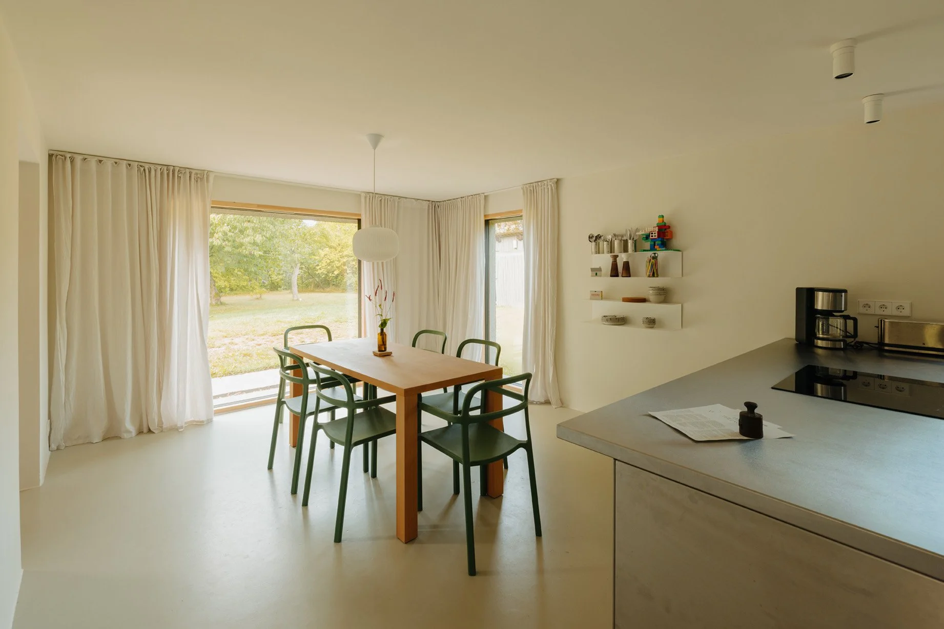 Bright dining room with wooden table and six green chairs, large windows with white curtains, view of outdoors, white walls, kitchen counter with coffee maker, and wall shelves with dishes and decorative items.