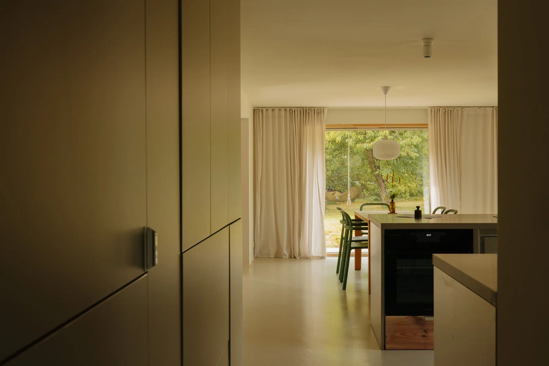 Mellenau 9. View of a modern dining area with a large window, green chairs around a table, and a hanging white light fixture, with curtains and a backyard visible outside.