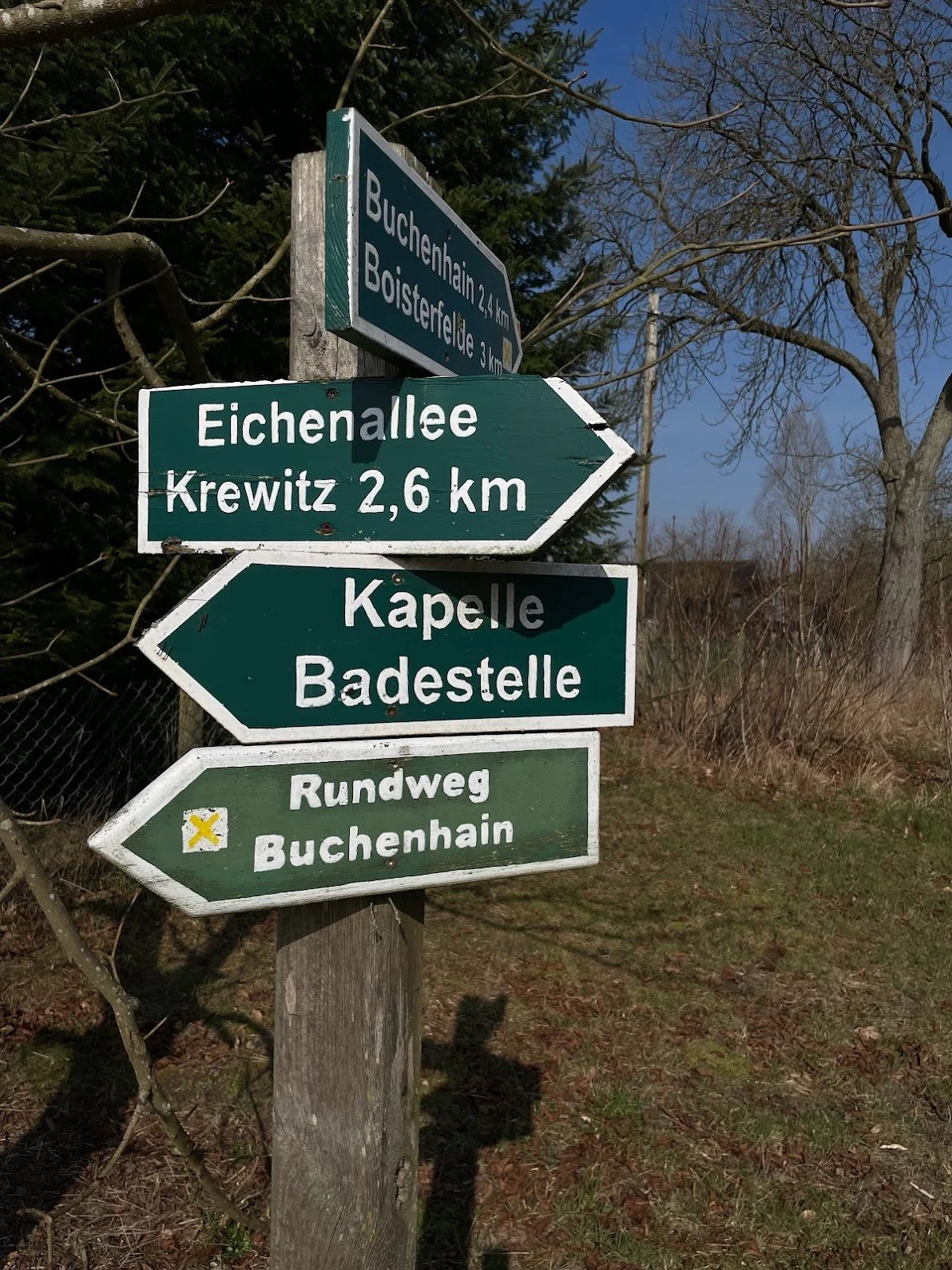 A wooden trail signpost with multiple green and blue directional signs with white text, indicating distances to various locations, situated outdoors with trees and a blue sky in the background.
