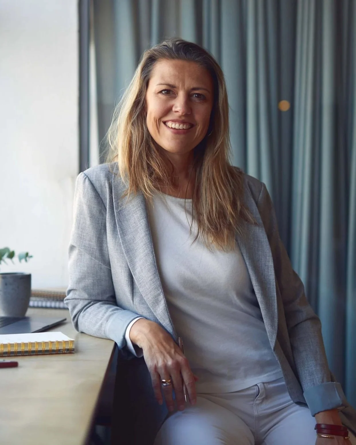 Kvindelig person sidder ved et bord i et kontorlokale, smilende, med bleg blazer og hvid top, der er omgivet af kontorartikler og planter.