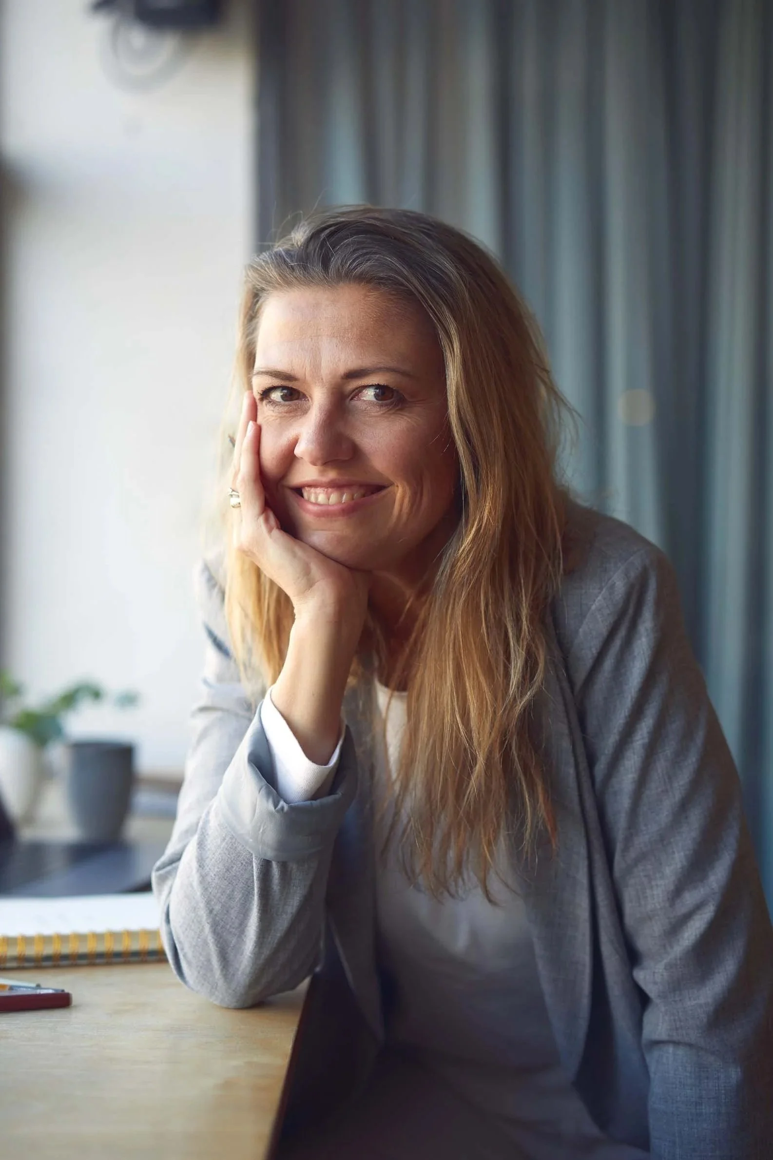 Kvindelig person med langt, lyst hår, sidder ved et bord med hånd under hage, smilende, i et hyggeligt, indendørs miljø med baggrund af blå gardiner.