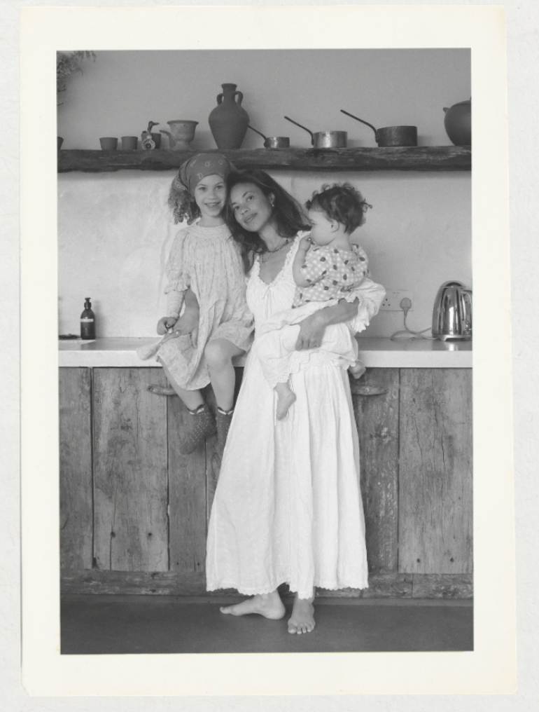 A woman holding a young girl and a toddler in her arms in a rustic kitchen with wooden cabinets and ceramic dishes on a shelf.