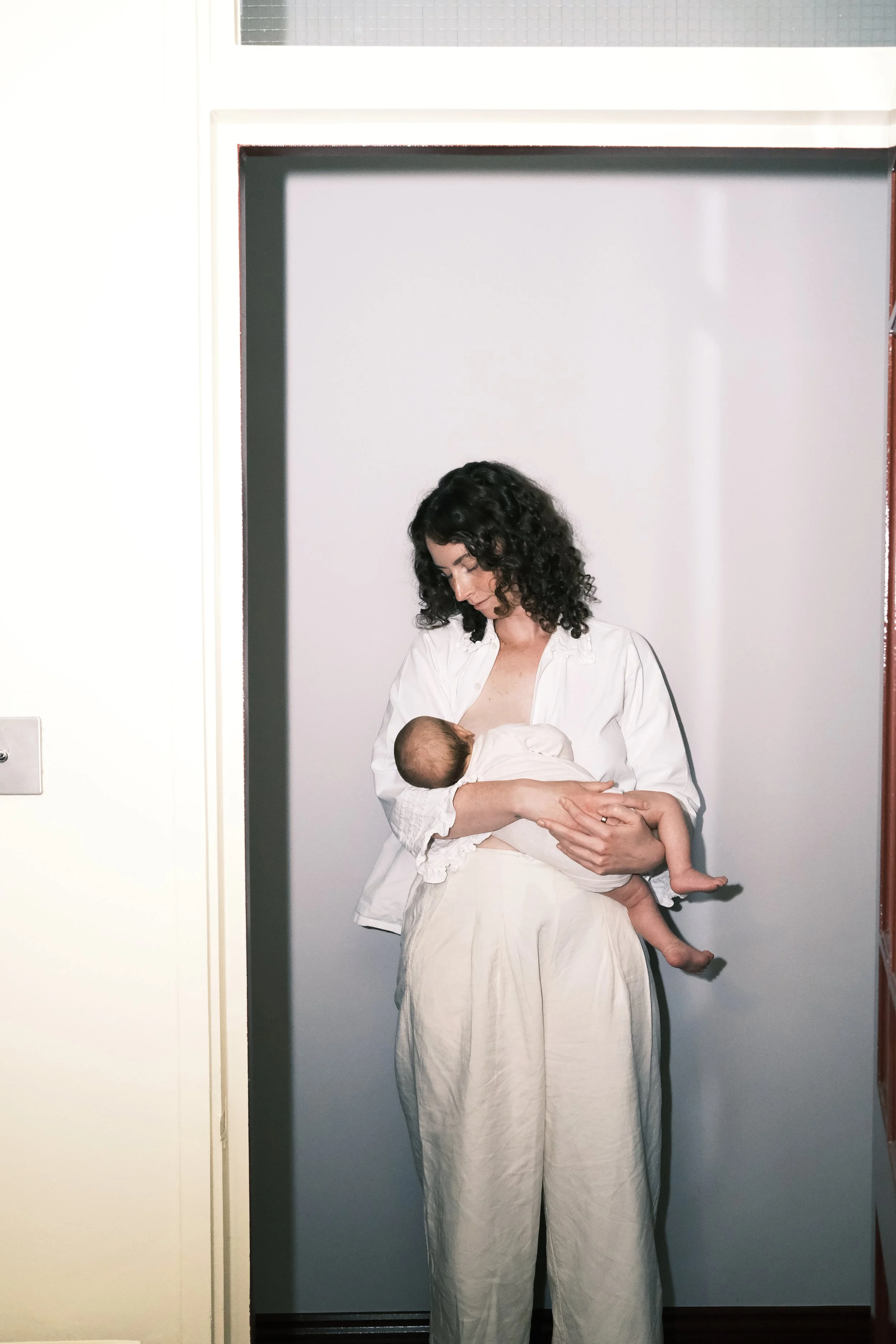 A woman with curly dark hair wearing a white shirt and white pants holds a sleeping baby in her arms. They are standing in front of a plain wall, with part of an open door visible.